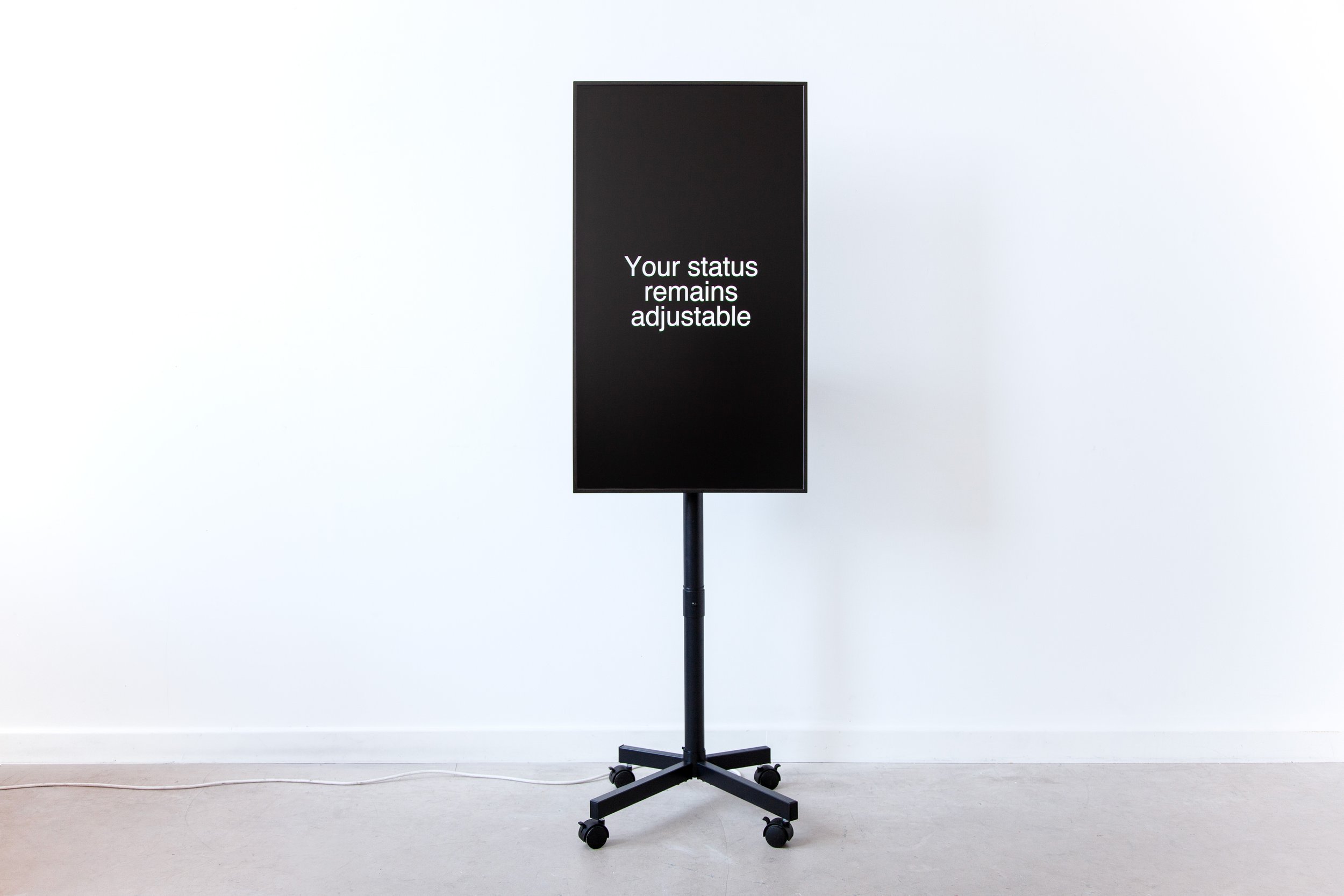 A tall black rectangular screen mounted on a wheeled black stand, displayed in a white gallery space with a concrete floor. The screen shows the phrase "Your status remains adjustable" in white text centered on a black background.