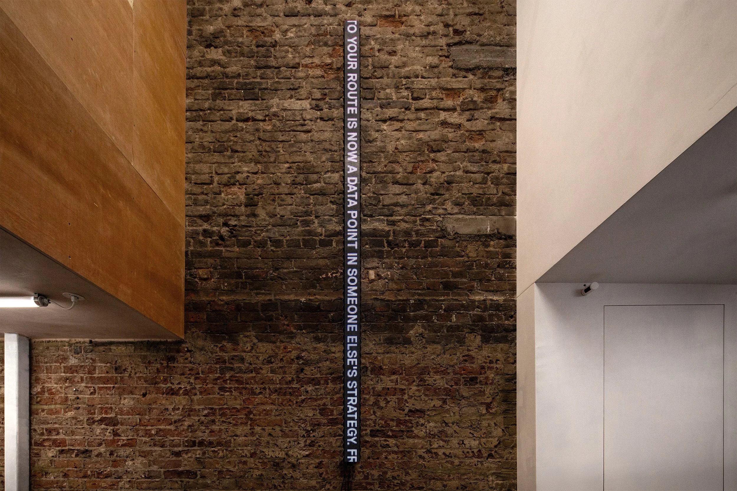 A narrow vertical LED ticker mounted on an exposed brick wall in a converted industrial interior, displaying the rotated text 'TO YOUR ROUTE IS NOW A DATA POINT IN SOMEONE ELSE'S STRATEGY.