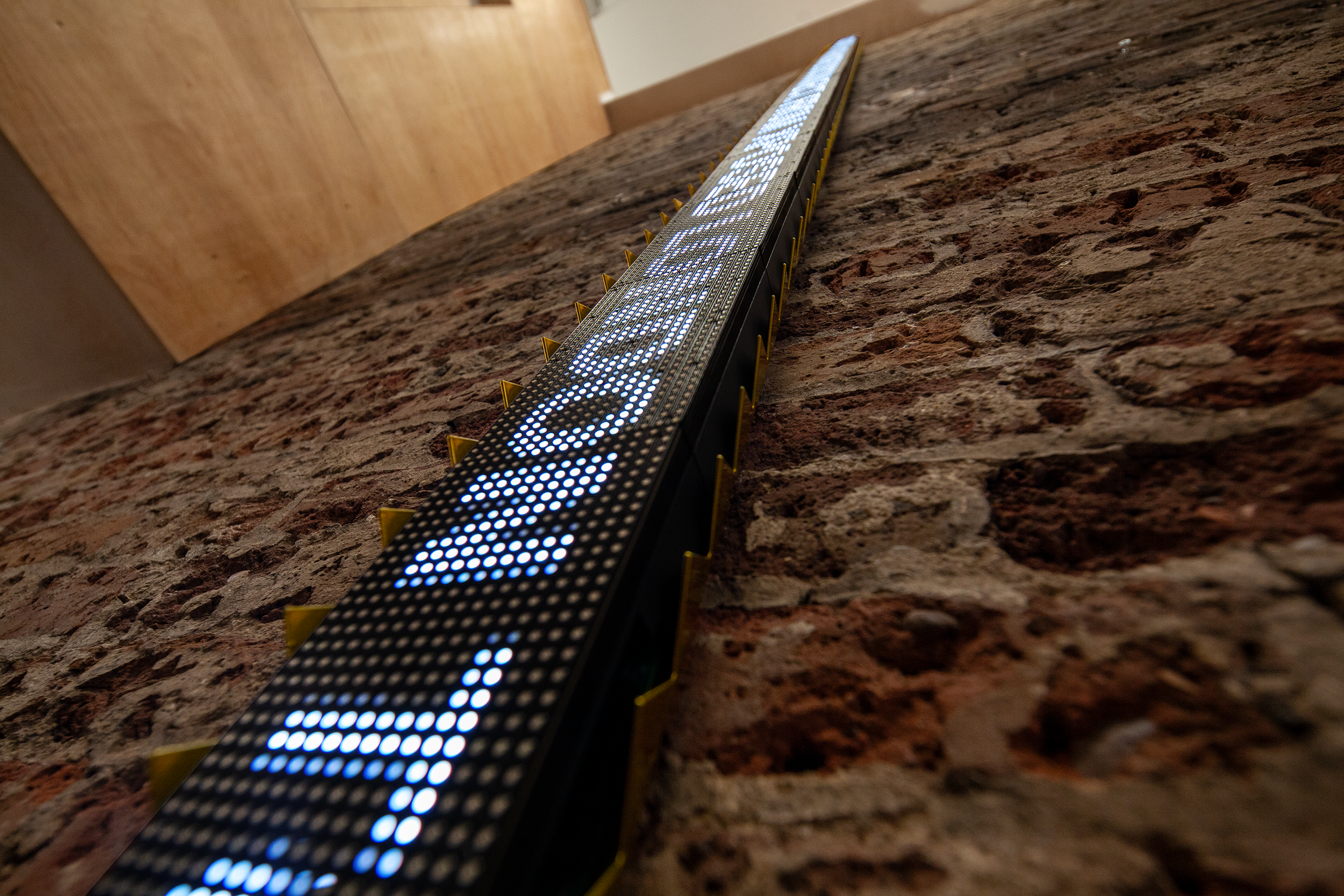 A narrow vertical LED ticker mounted on an exposed brick wall in a converted industrial interior, displaying rotated text, the display is encased in security spikes powder coated in gold