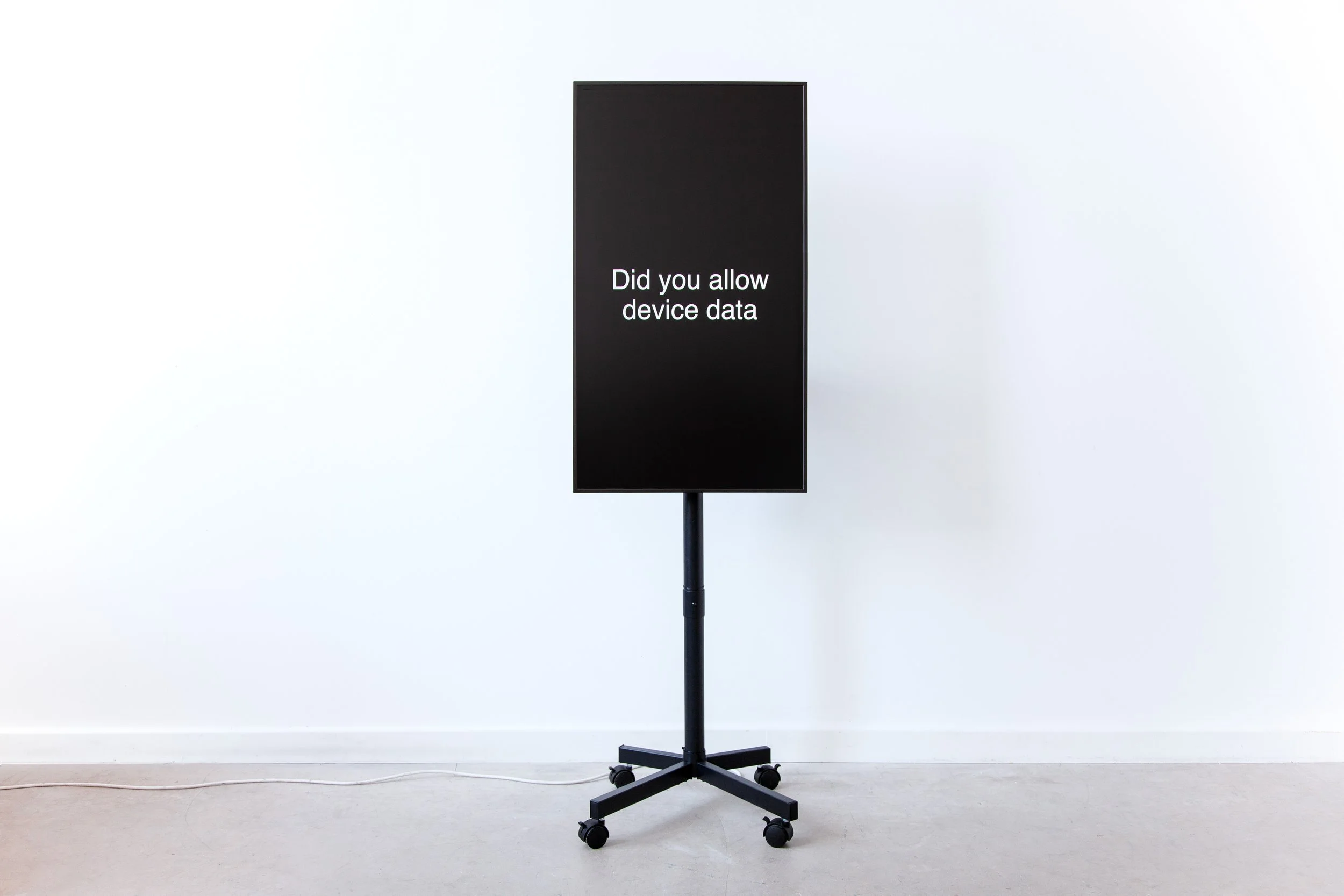 A tall black rectangular screen mounted on a wheeled black stand, displayed in a white gallery space with a concrete floor. The screen shows the phrase "Did you allow device data" in white text centered on a black background.