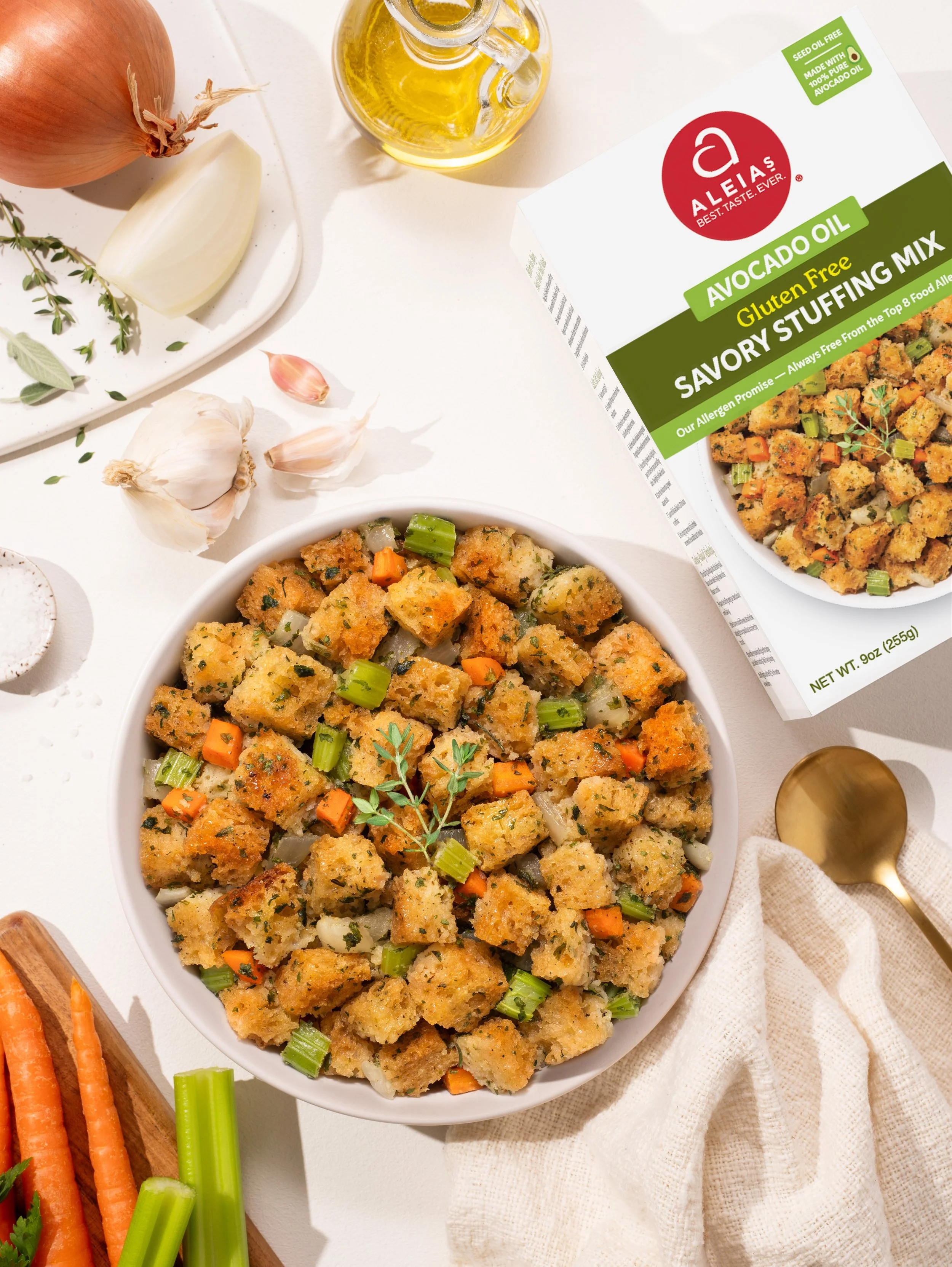 A bowl of gluten-free avocado stuffing mix with bread cubes, chopped celery and carrots, garnished with fresh herbs, placed on a white surface with garlic, onion, and garlic bulb nearby, and a box of Altaia savory stuffing mix in the background.