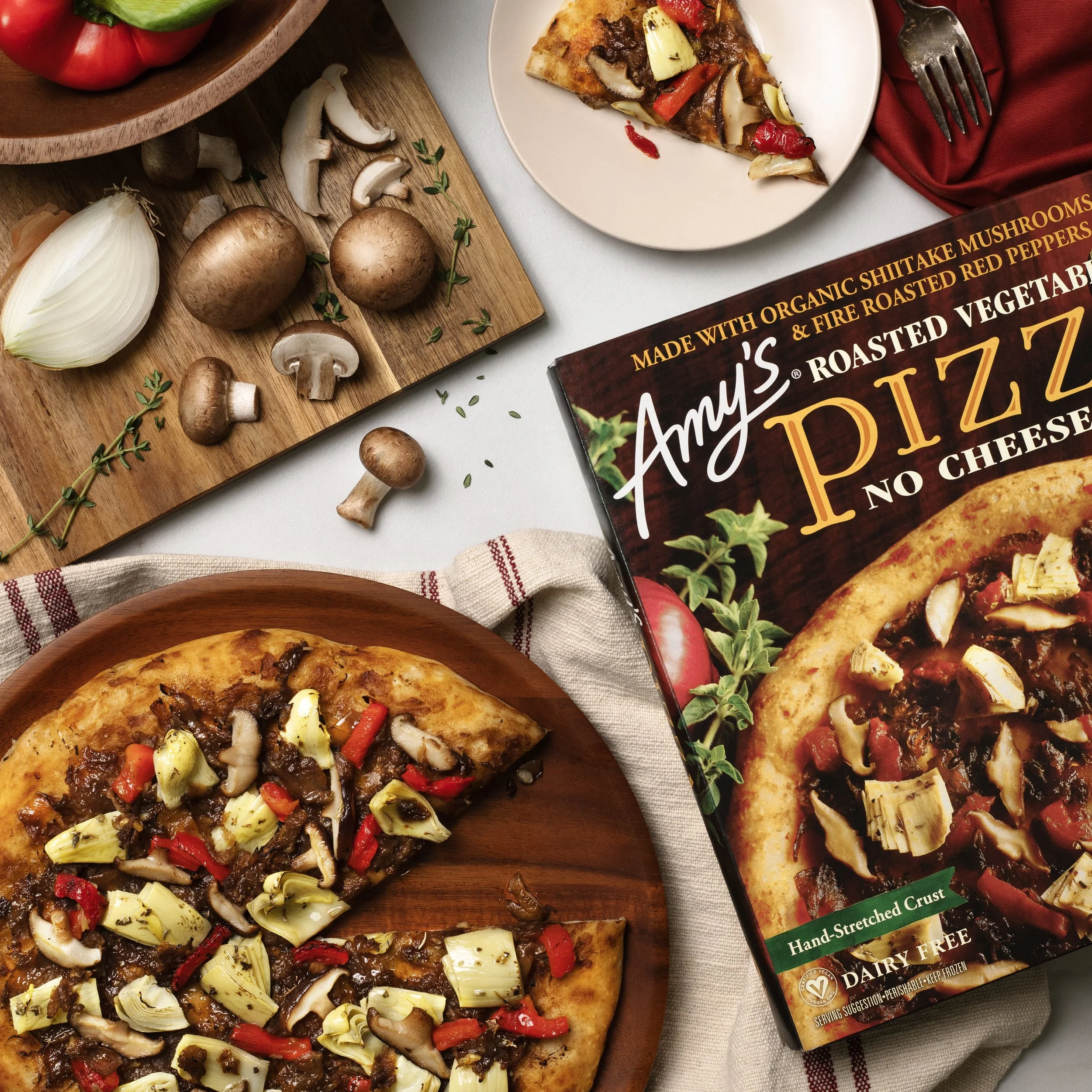 A vegetarian frozen pizza topped with artichokes, mushrooms, roasted red peppers, and cheese, with a box of Amy's organic mushroom pizza nearby, a wooden cutting board with fresh mushrooms, garlic, and herbs, and a plate with a slice of pizza on a white surface.