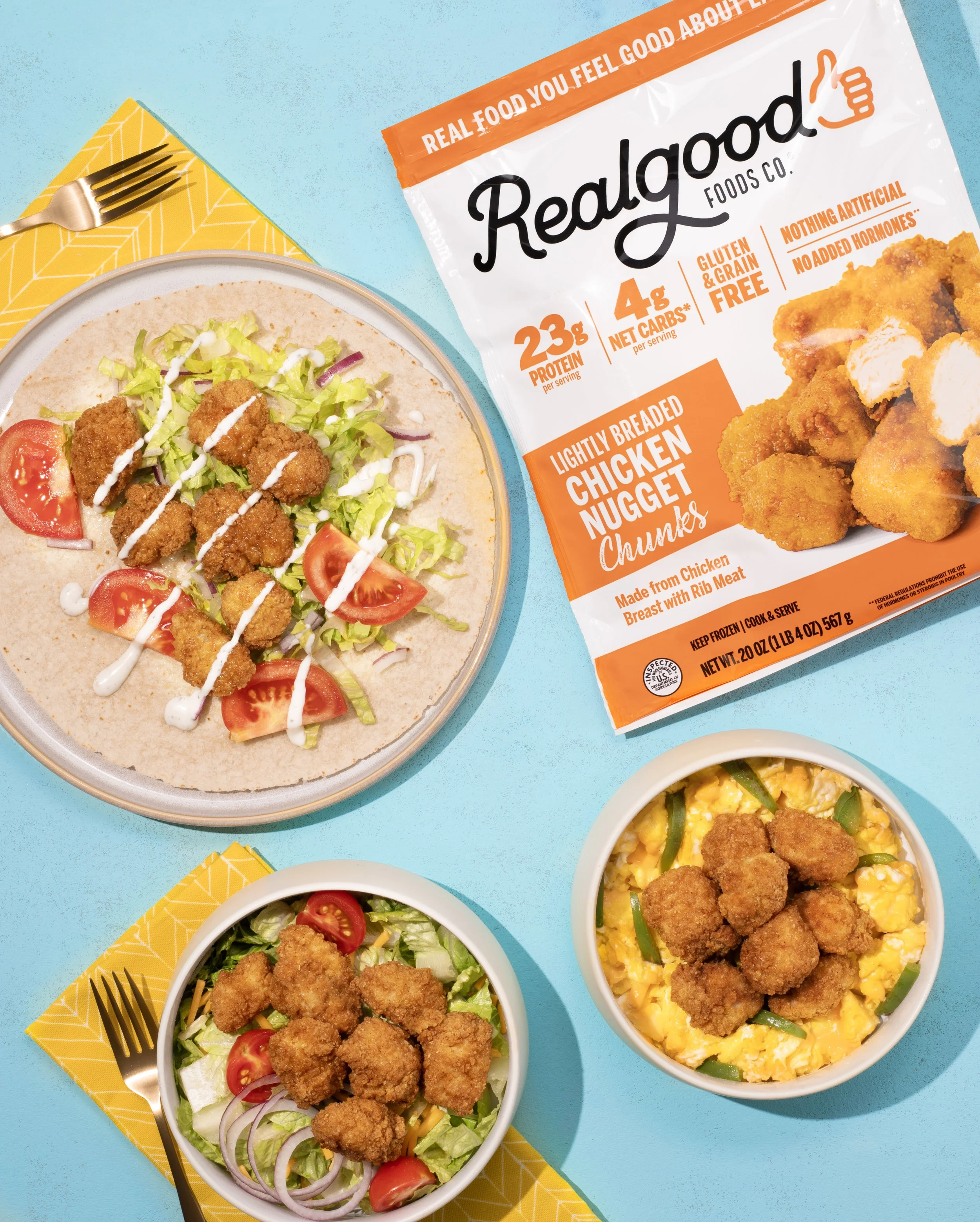 A plate with a taco containing fried chicken chunks, lettuce, tomato, and drizzled sauce; two bowls with fried chicken chunks over salad and scrambled eggs with vegetables; a package of Lightly Breaded Chicken Nugget Chunks from Real Good Foods on a blue surface with yellow napkins and gold forks.