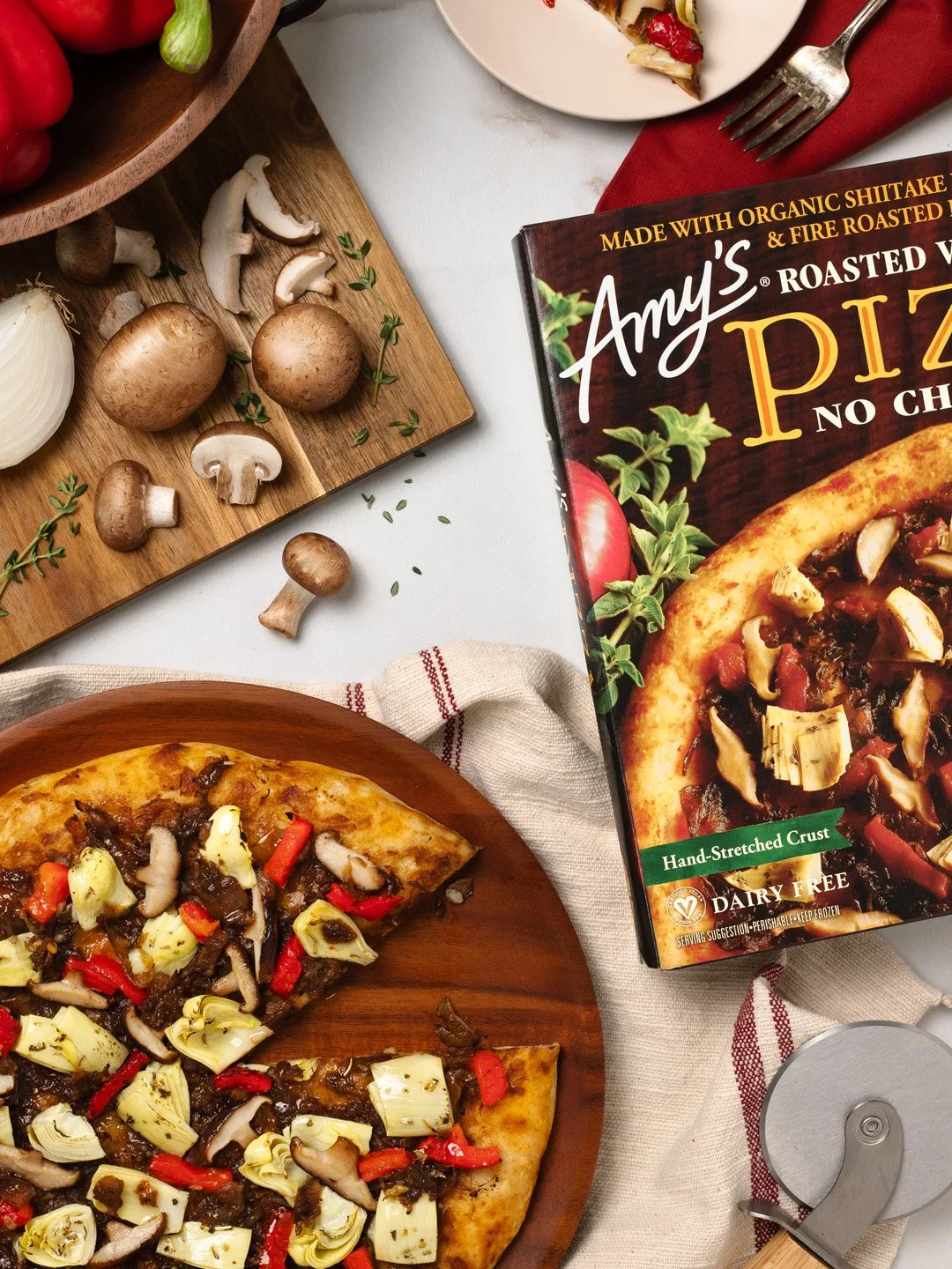 Vegetarian mushroom pizza with artichokes and red peppers on a wooden plate, mushrooms and garlic on a cutting board, Amy's vegan pizza box, and a pizza cutter.