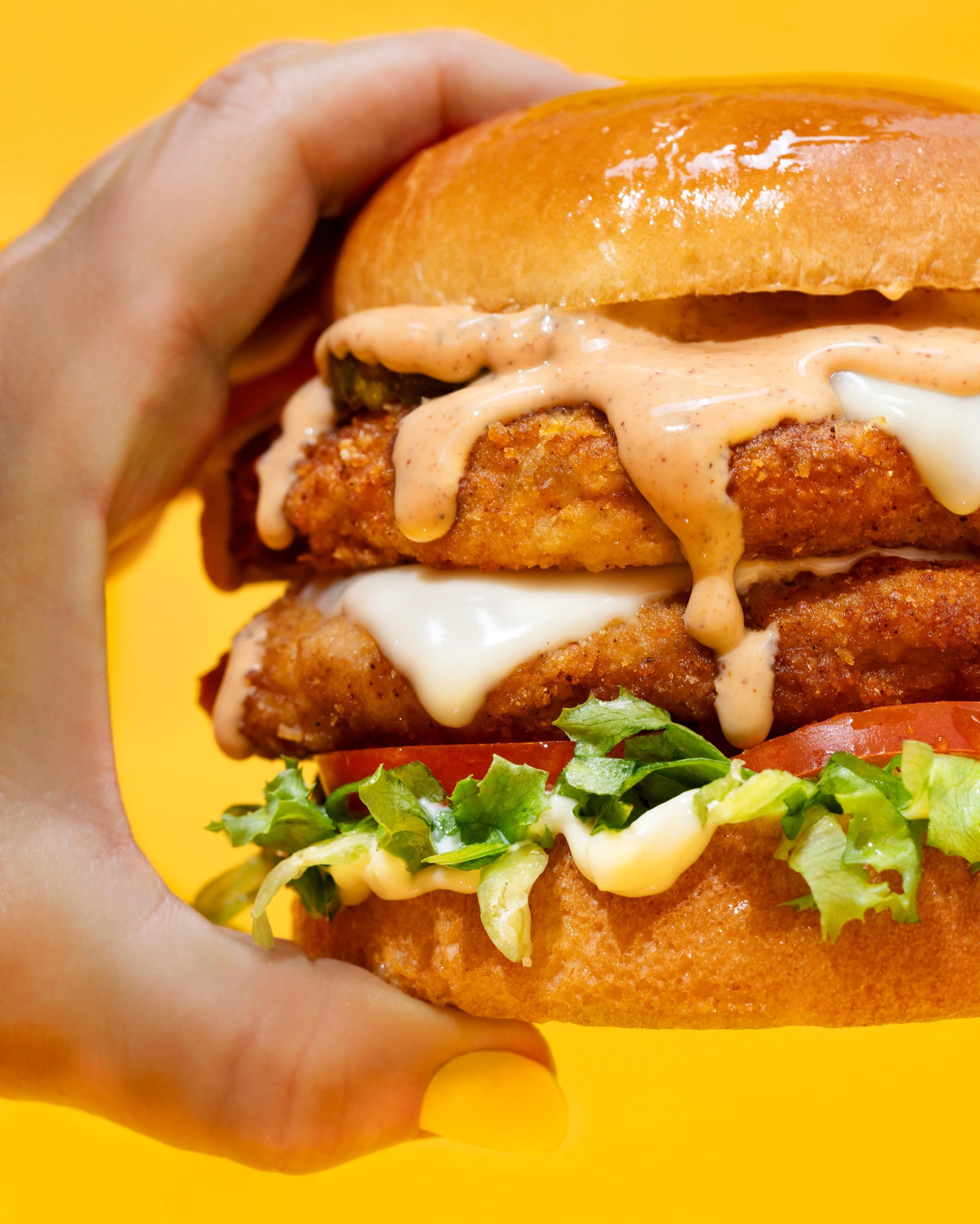 Close-up of a hand holding a crispy chicken sandwich with lettuce, tomato, and multiple sauces on a yellow background.