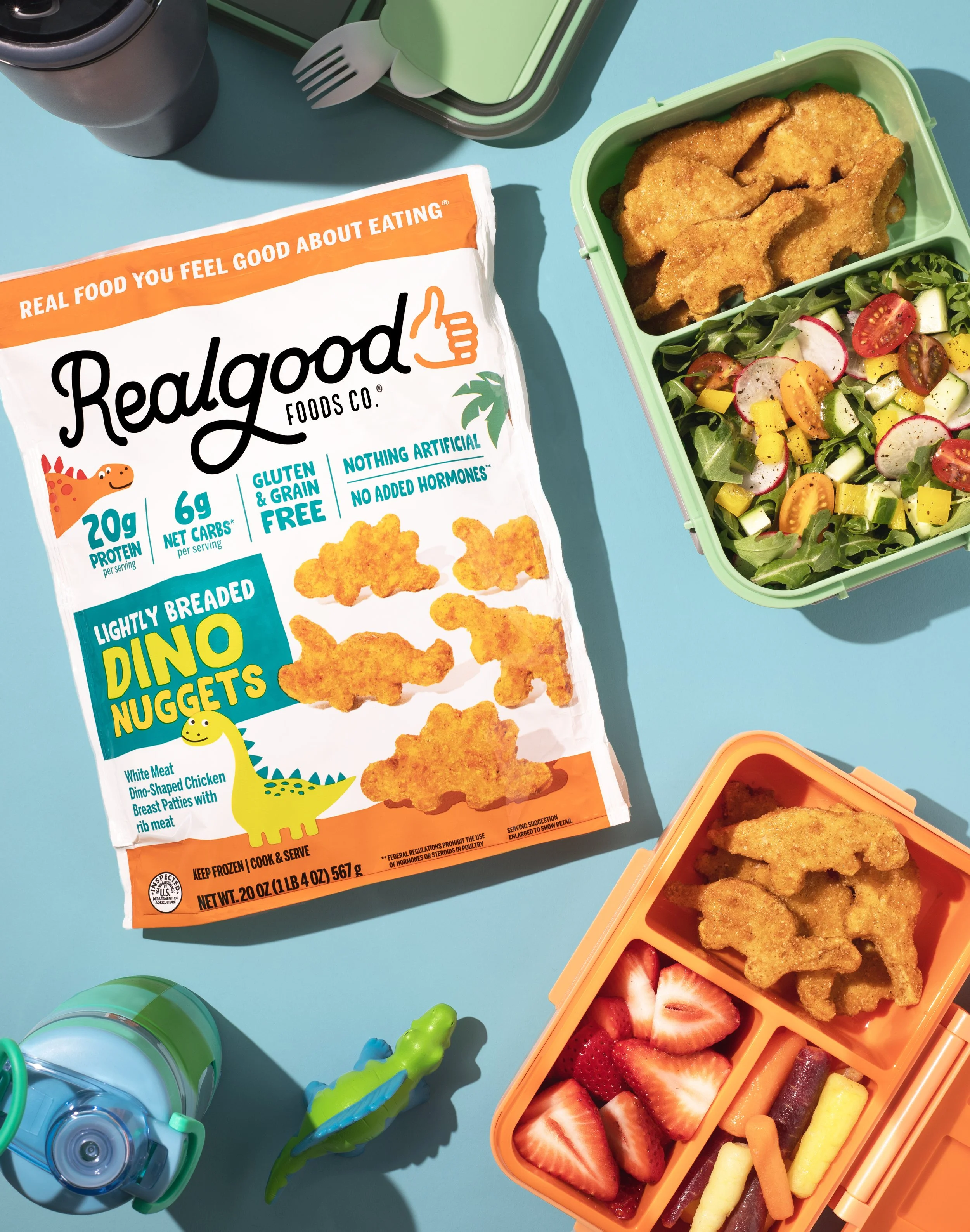 Packaged dinosaur-shaped chicken nuggets with a salad, strawberries, and lunch items on a blue background.