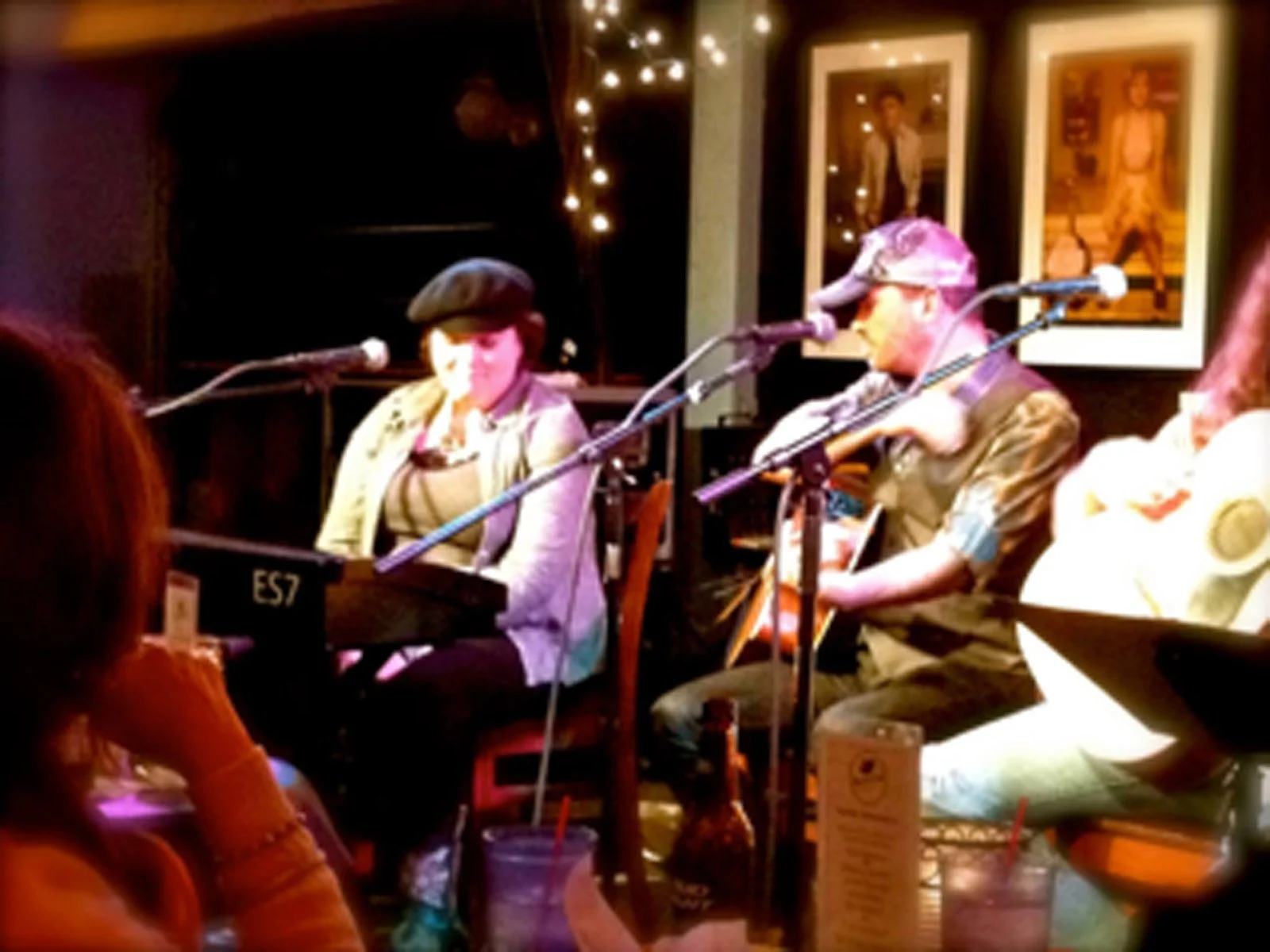 Two musicians performing on stage at a cozy venue, one playing keyboard and the other playing guitar and singing, with framed photos on the wall behind them and string lights overhead.