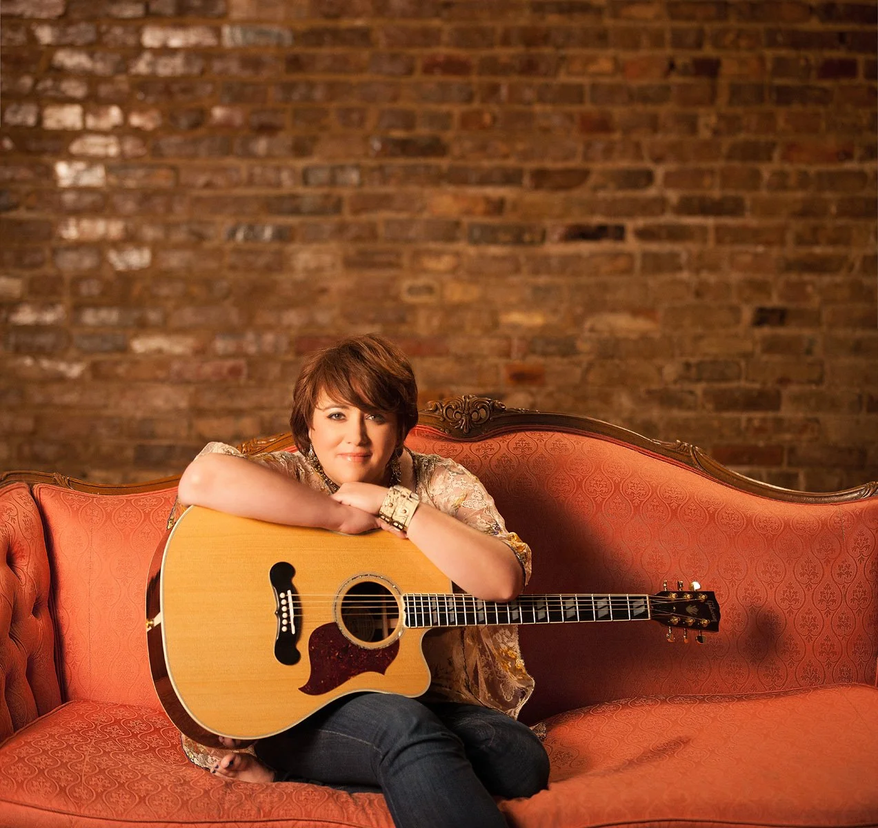 A woman sitting on an orange vintage sofa, holding an acoustic guitar, with a brick wall background.