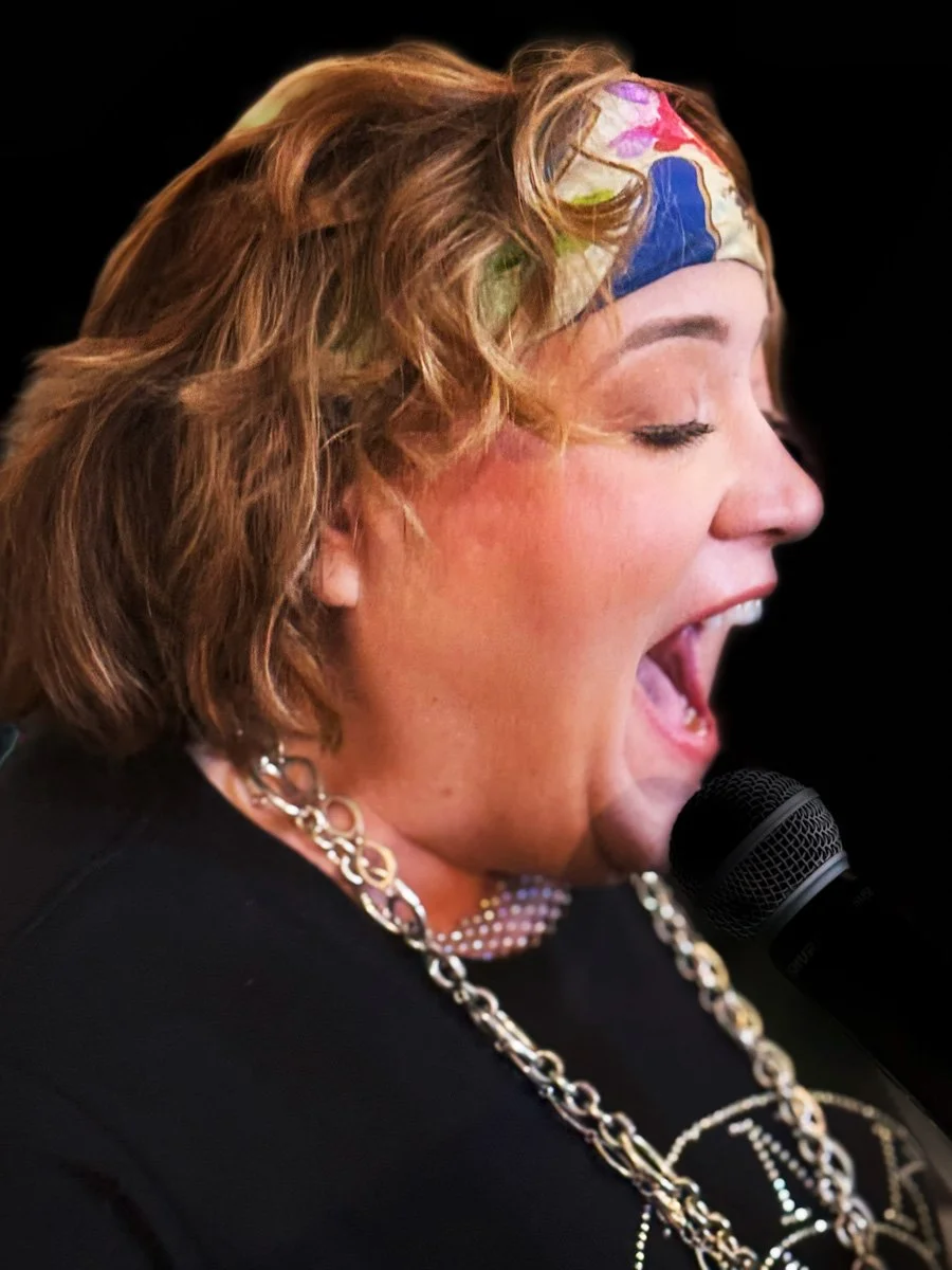 A woman with short, wavy red hair wearing a colorful headscarf, earrings, and layered necklaces, passionately speaking or singing into a microphone, with her mouth open and eyes closed, against a black background.