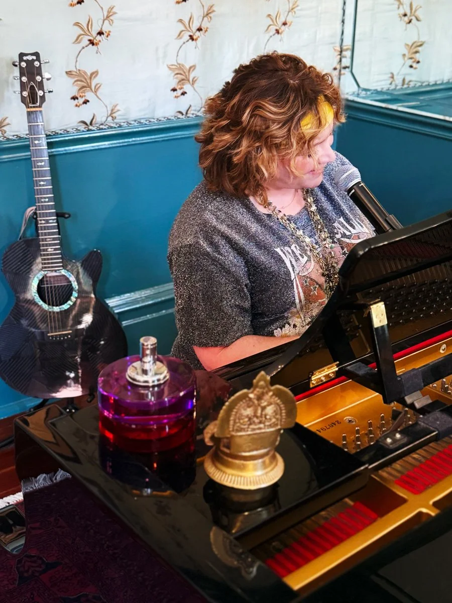 A woman with curly brown hair wearing a dark sparkly shirt, singing into a microphone while playing a grand piano. A black acoustic guitar leans against the wall in the background.