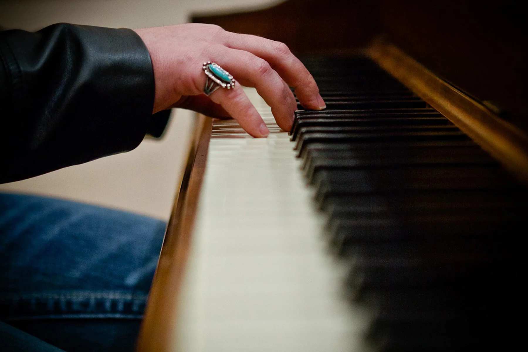 A person playing a piano, focusing on their hand with a large turquoise and silver ring, wearing a black leather jacket, on the piano keys.