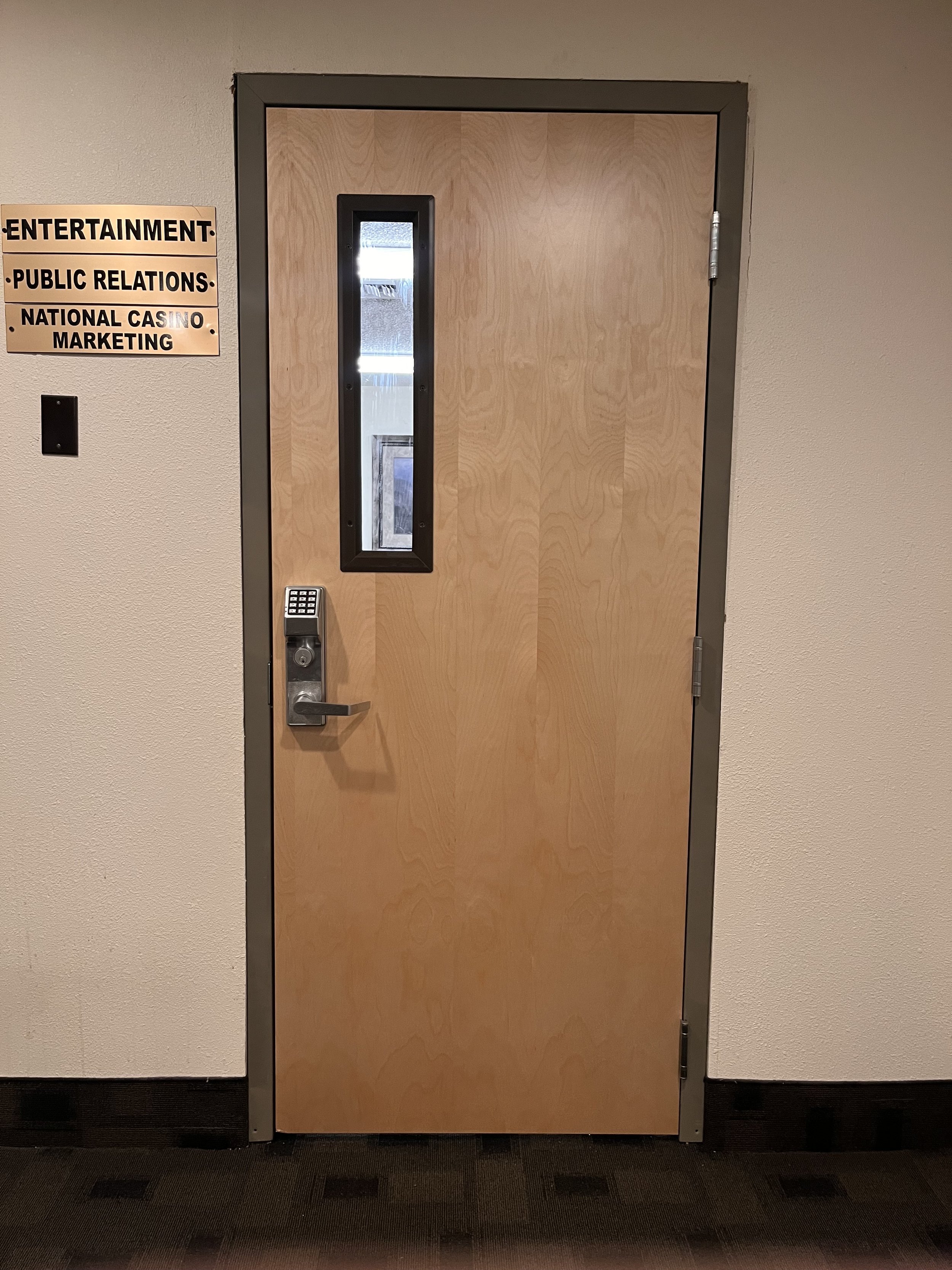 Wooden door with a small vertical window, electronic keypad lock, and door handle. Signs on the wall to the left with rules and restrictions.