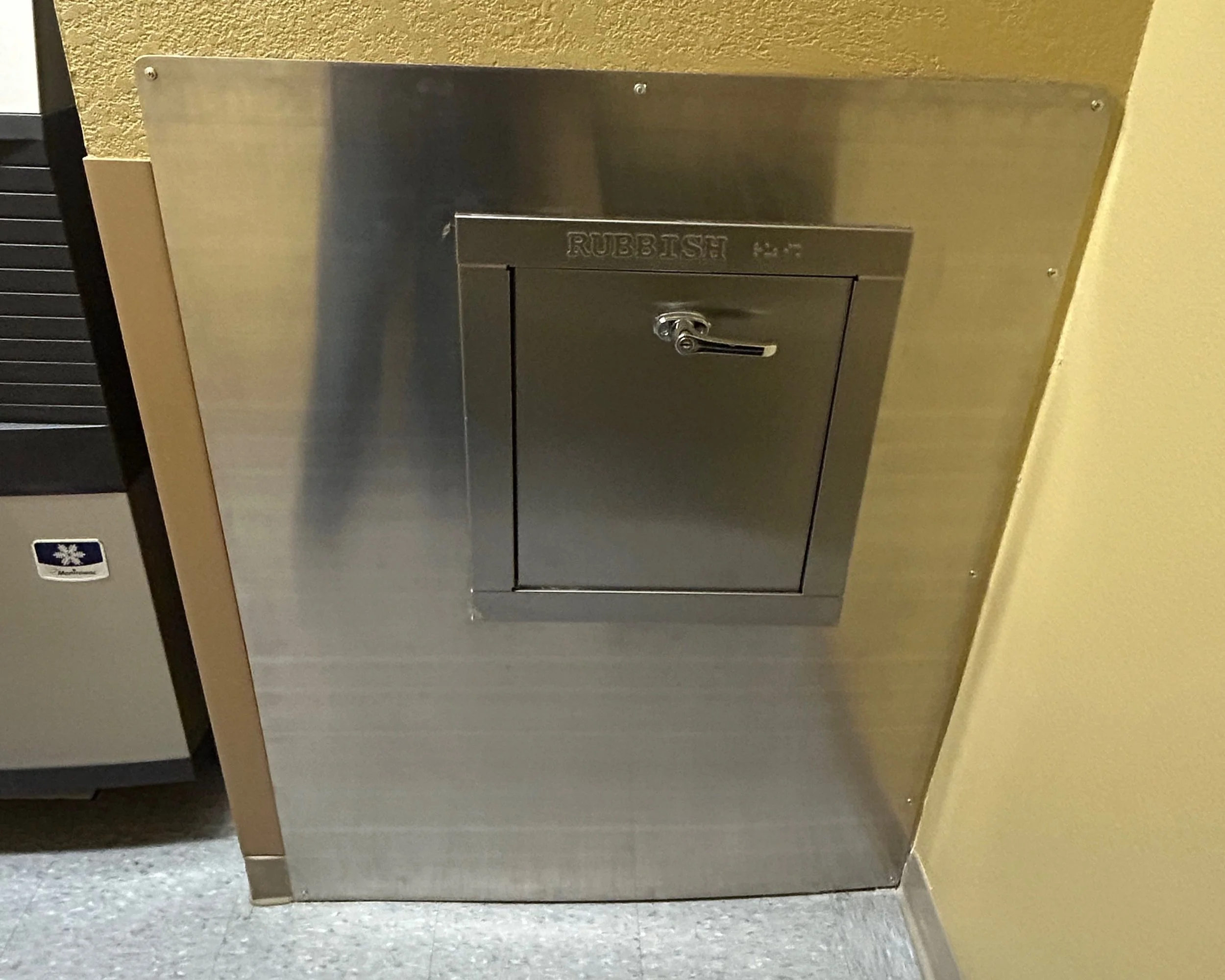 A metal rubbish chute door labeled 'RUBBISH' with a handle, built into a wall in a building interior.