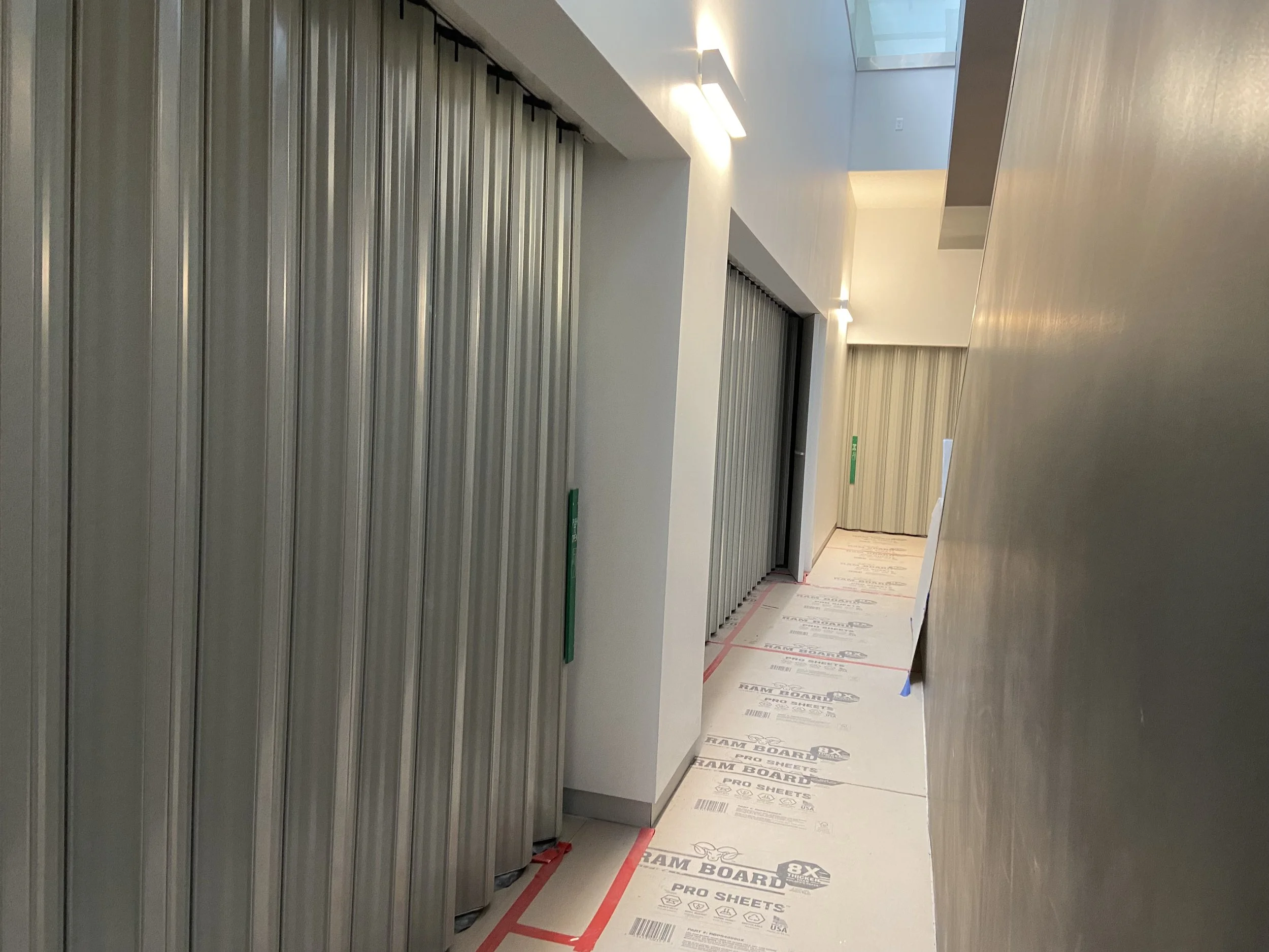 Hallway with metal roll-up doors on the left, white walls with built-in lights, and temporary flooring covering the floor during construction.
