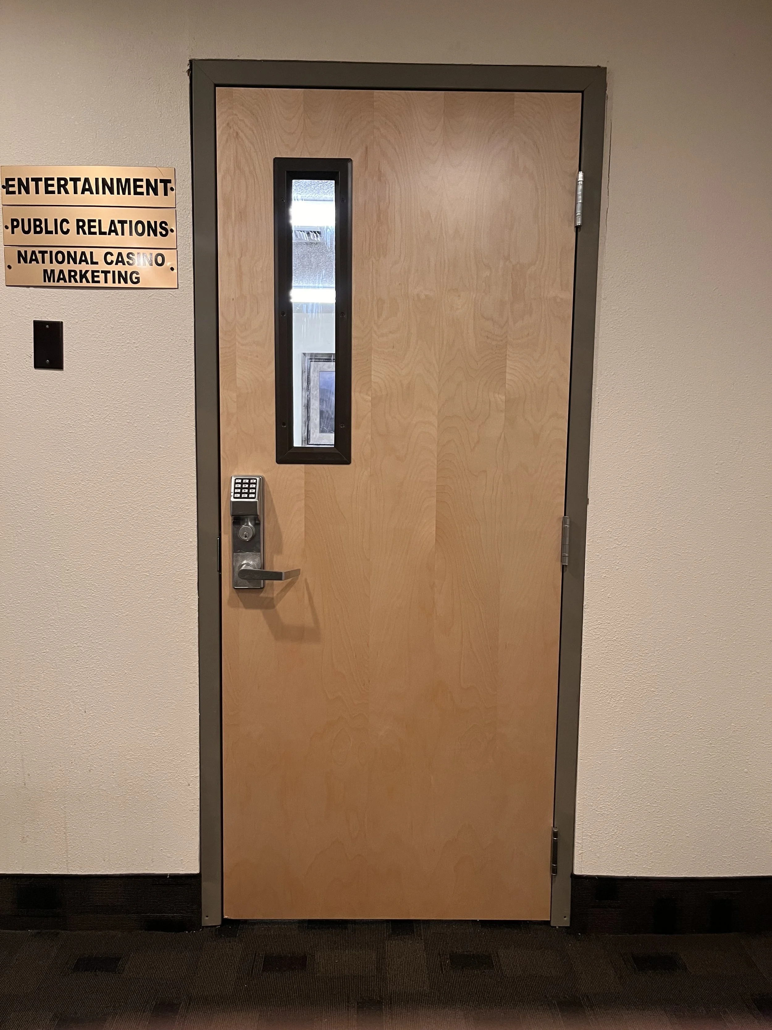 A closed wooden door with a small vertical window, an electronic keypad lock, and a lever handle, set in a beige wall with a sign nearby.
