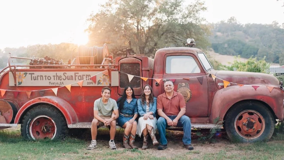 Four people sitting in front of an old fire truck decorated with a banner that reads "Turn to the Sun Farm, Penryn, CA, 2014," on a grassy field during sunset with trees and hills in the background.