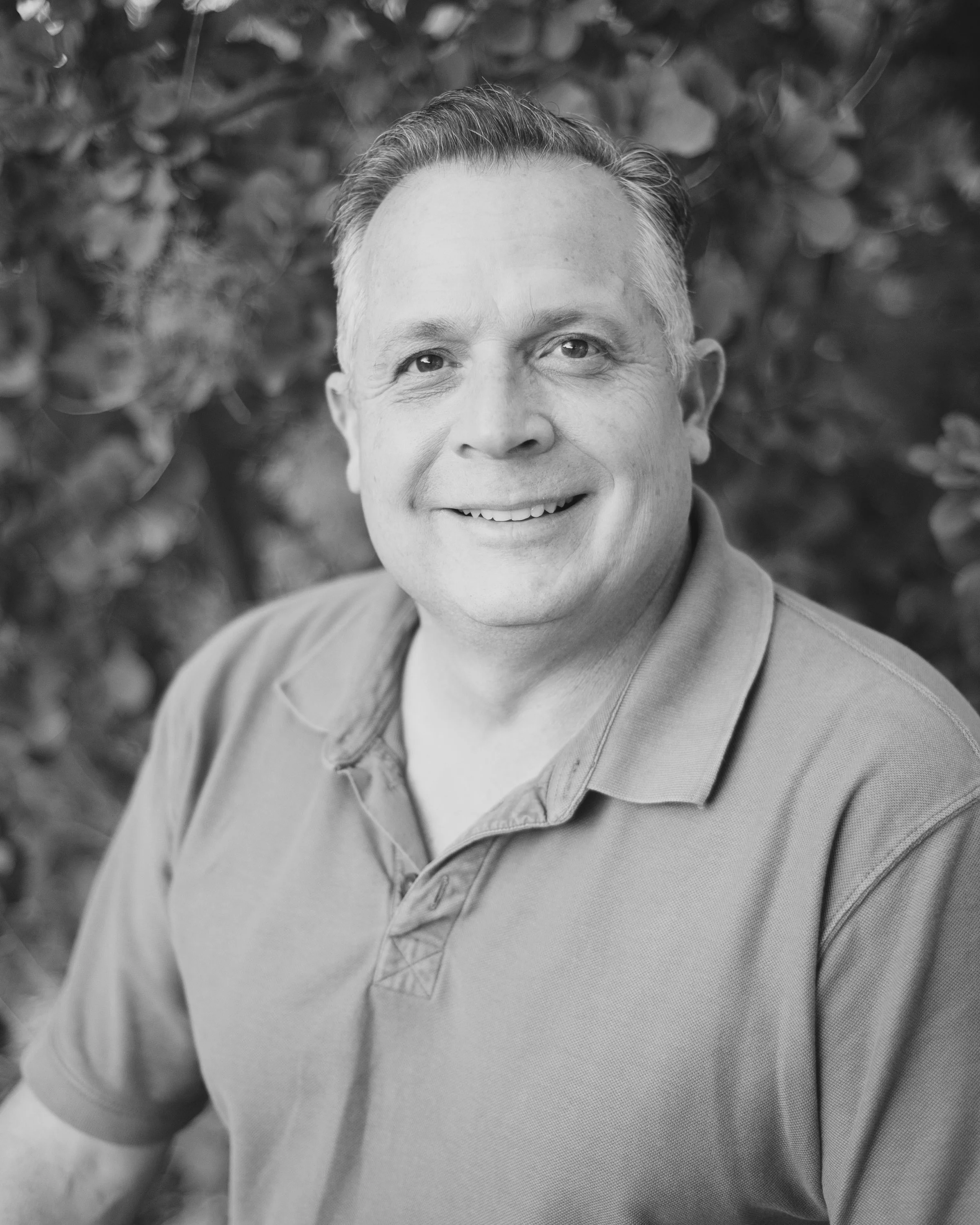 A smiling middle-aged man with short hair, wearing a polo shirt, outdoors with blurred foliage in the background.