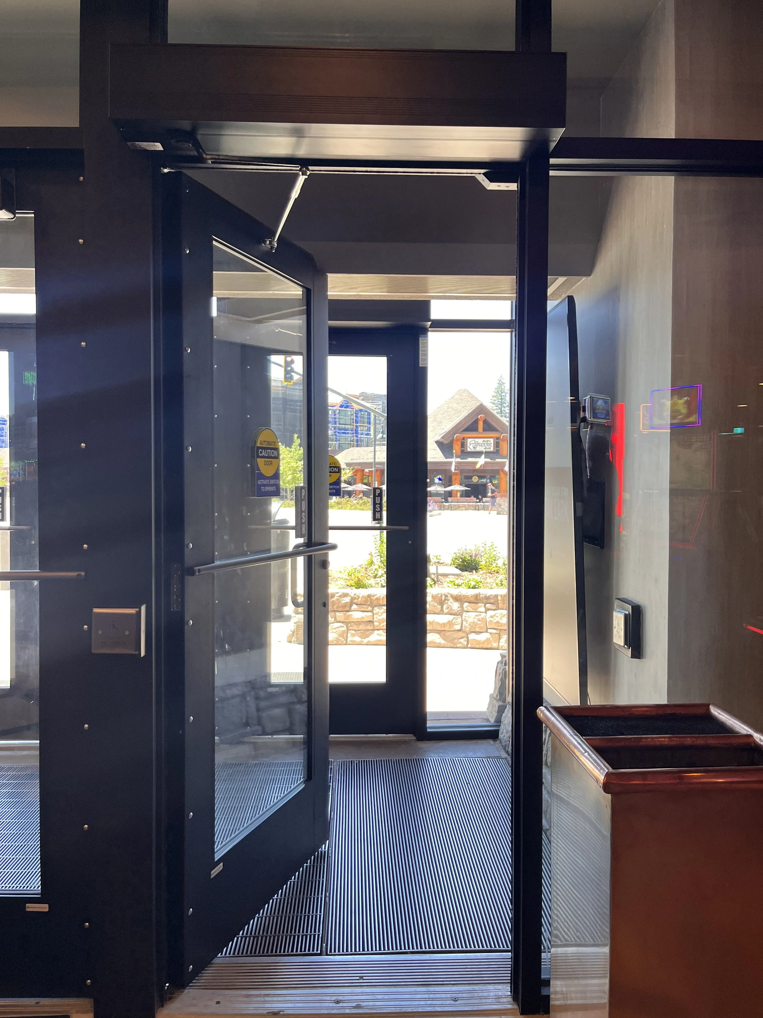Glass door with push bar and safety signs, view of shopping area exterior with buildings and trees.