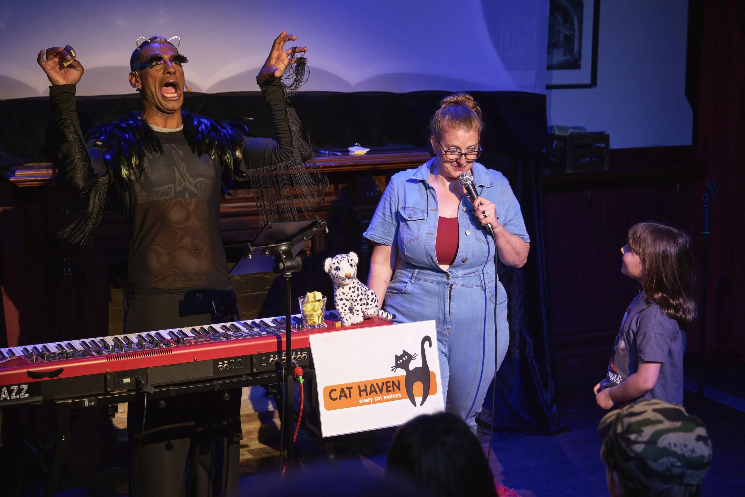 Comedian Robyn Perkins and Gavin Nicklette speaking into a microphone, a child looking up at her, q display with 'Cat Haven' on it, stuffed tiger, and keyboard on stage.
