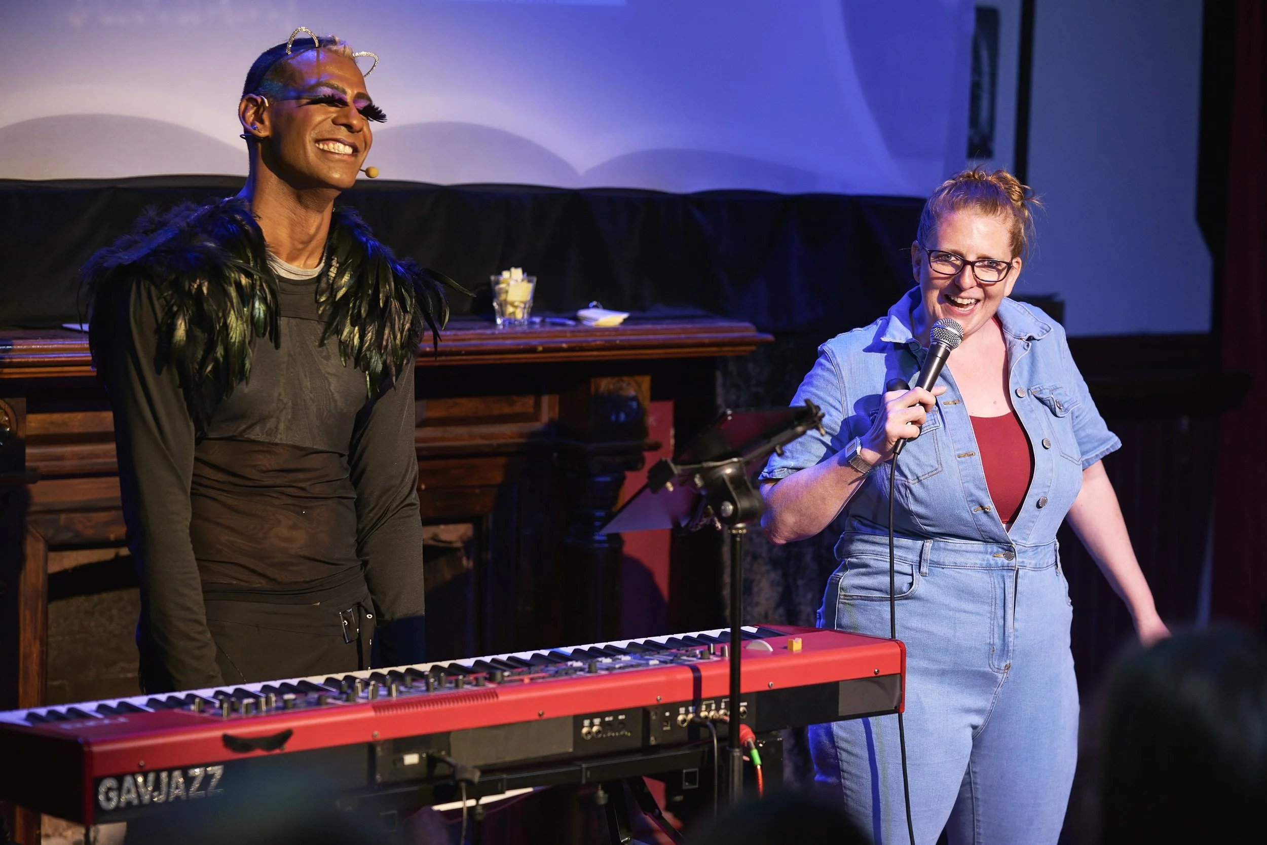 Comedian Robyn Perkins and Gavin Nicklette, one smiling with a microphone, the other standing near a red keyboard, in a dimly lit venue.