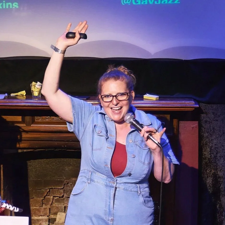 Comedian Robyn Perkins, holding a microphone and raising her right hand, is smiling and speaking on stage with a dark background and a bookshelf behind her.