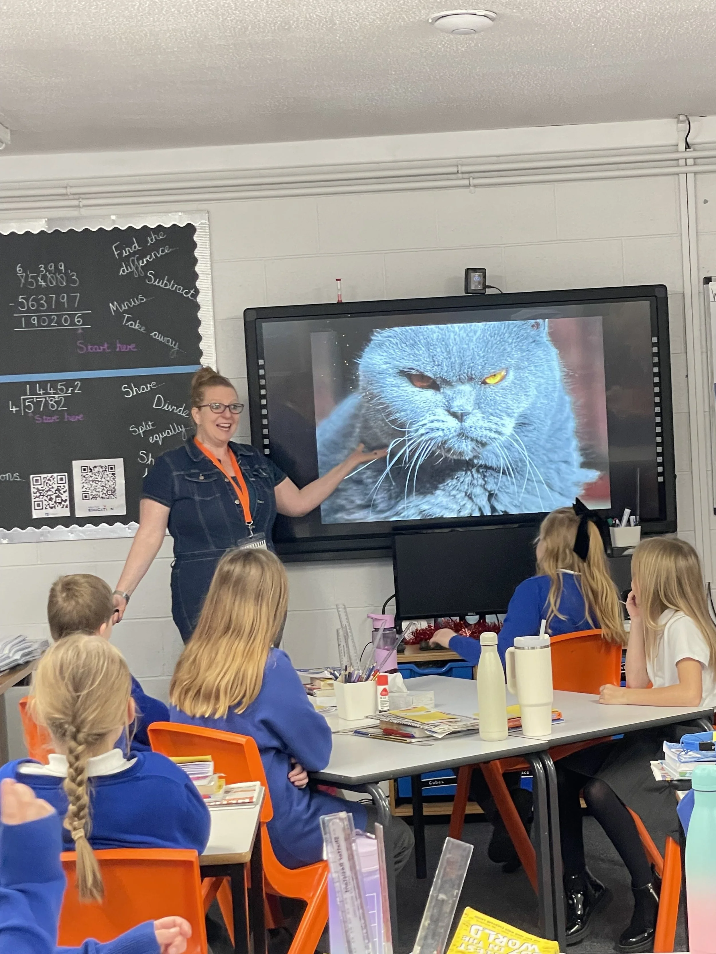 Comedian Robyn Perkins in a classroom pointing at a large screen showing a close-up of a grumpy grey cat with yellow and brown eyes while students observe. 