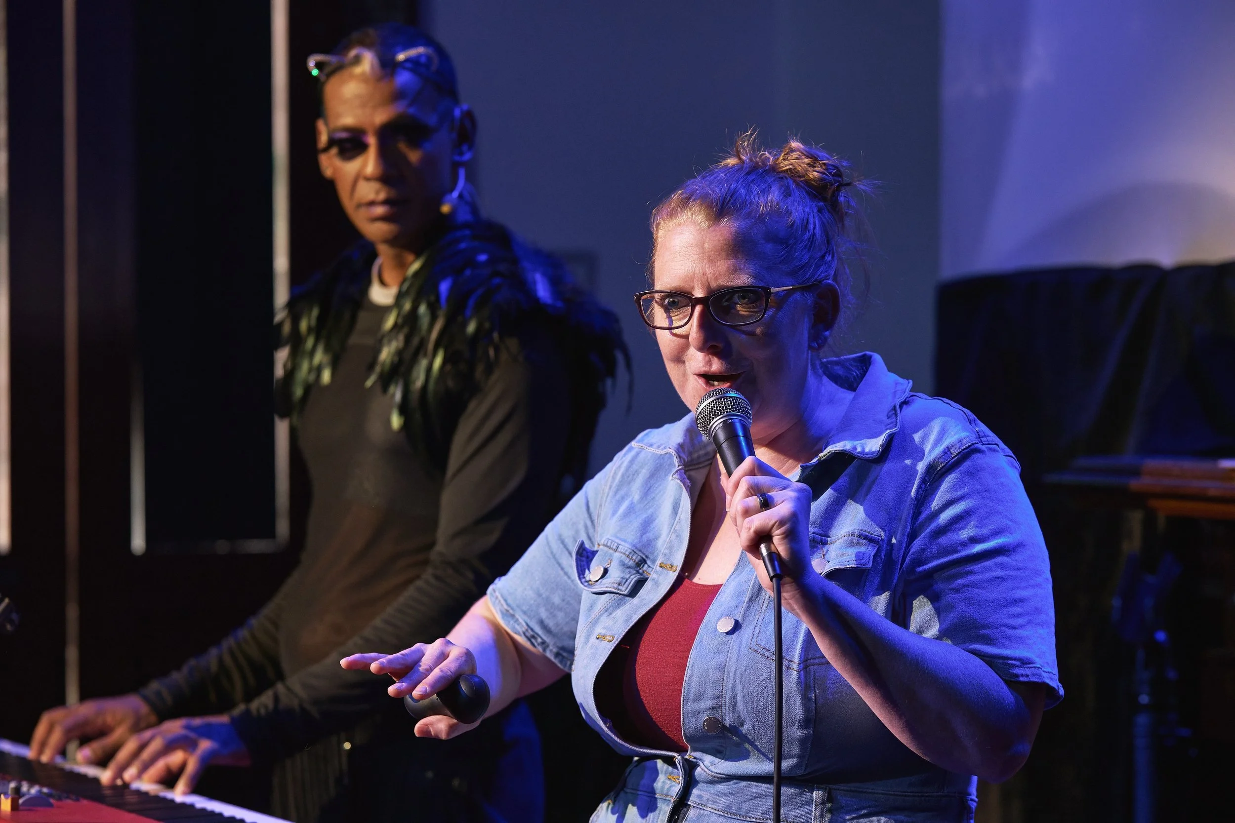 Comedian Robyn Perkins and Gavin Nicklette perfoming into a microphone and Gavin is playing a keyboard, wearing a black outfit with feathered shoulders.
