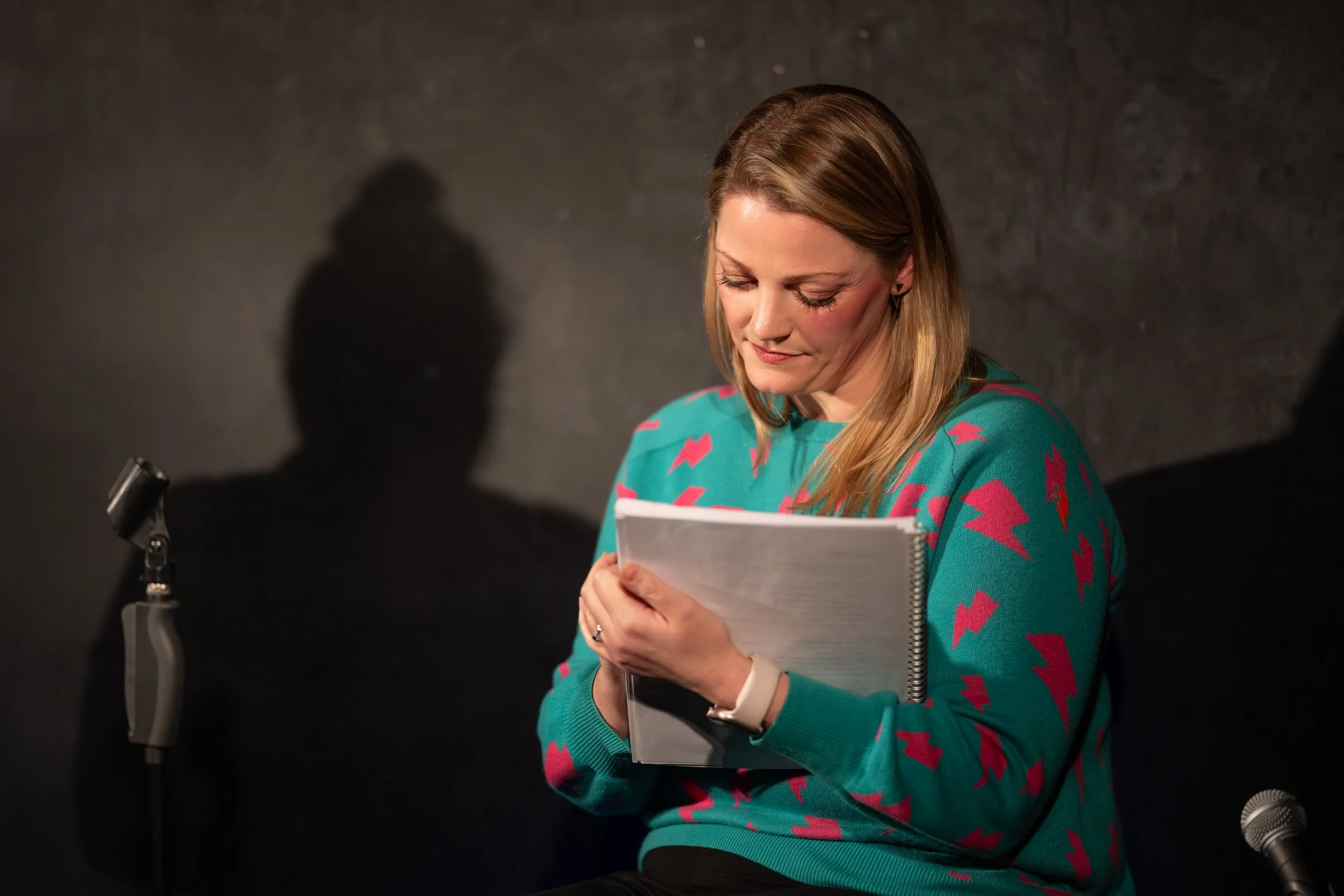 A woman with long blonde hair in a teal sweater with pink lightning bolts, holding a notebook, sitting in front of a dark background with a microphone and a stand shadow behind her.