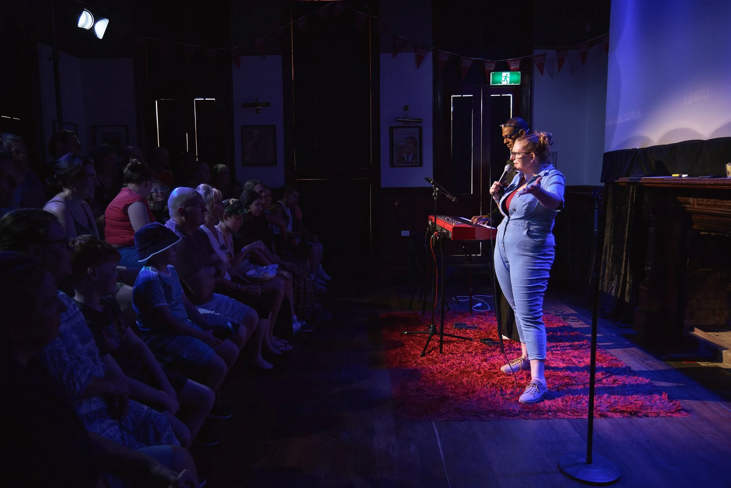 Comedian Robyn Perkins performing on stage with a microphone, audience seated watching, in a dimly lit room with colorful lighting and a red patterned rug.