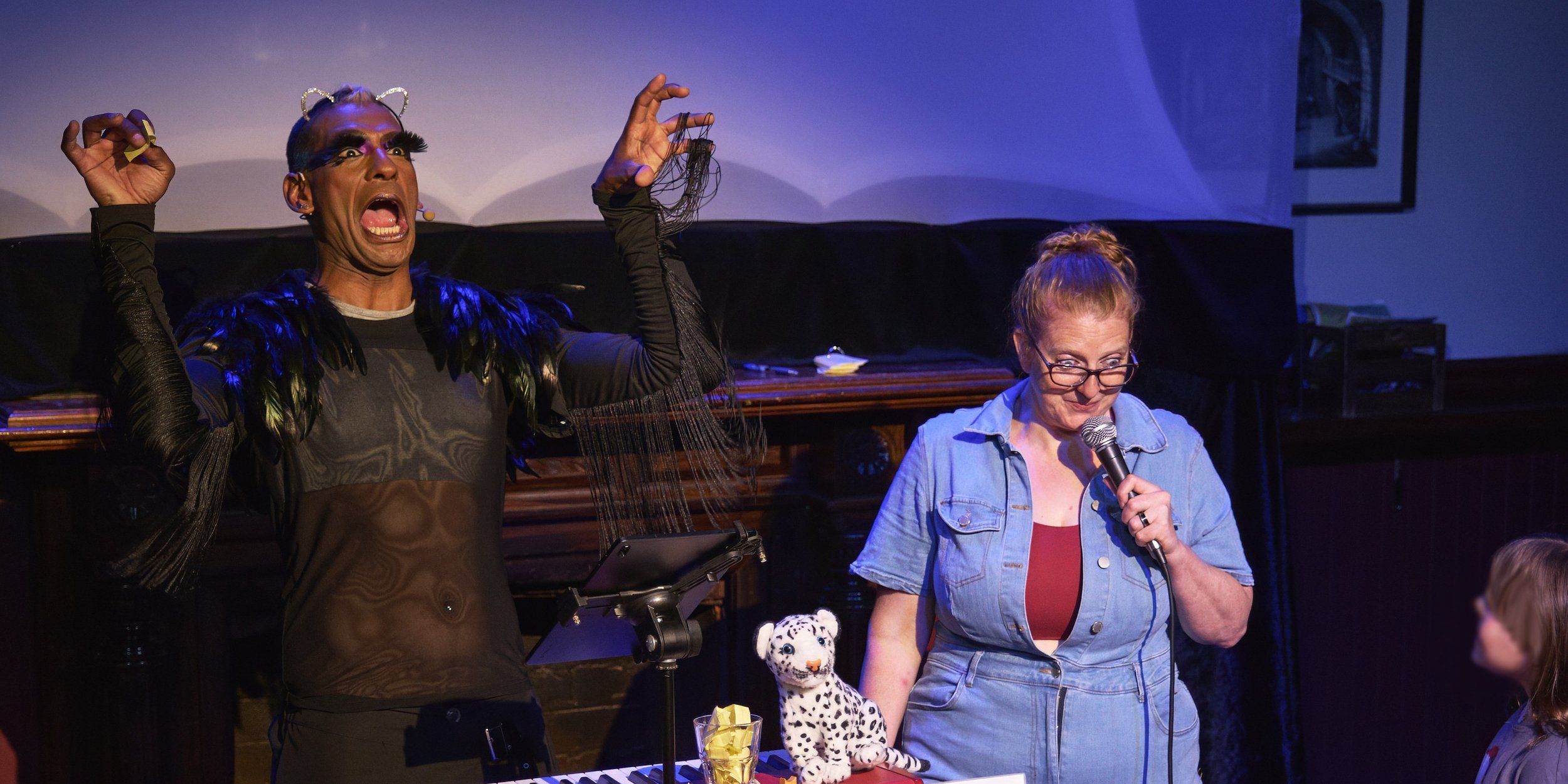 Comedian Robyn Perkins and Gavin Nicklette, holding a microphones and making an expressive gestures. They are on a stage with a piano, a stuffed cheetah, and a music stand.