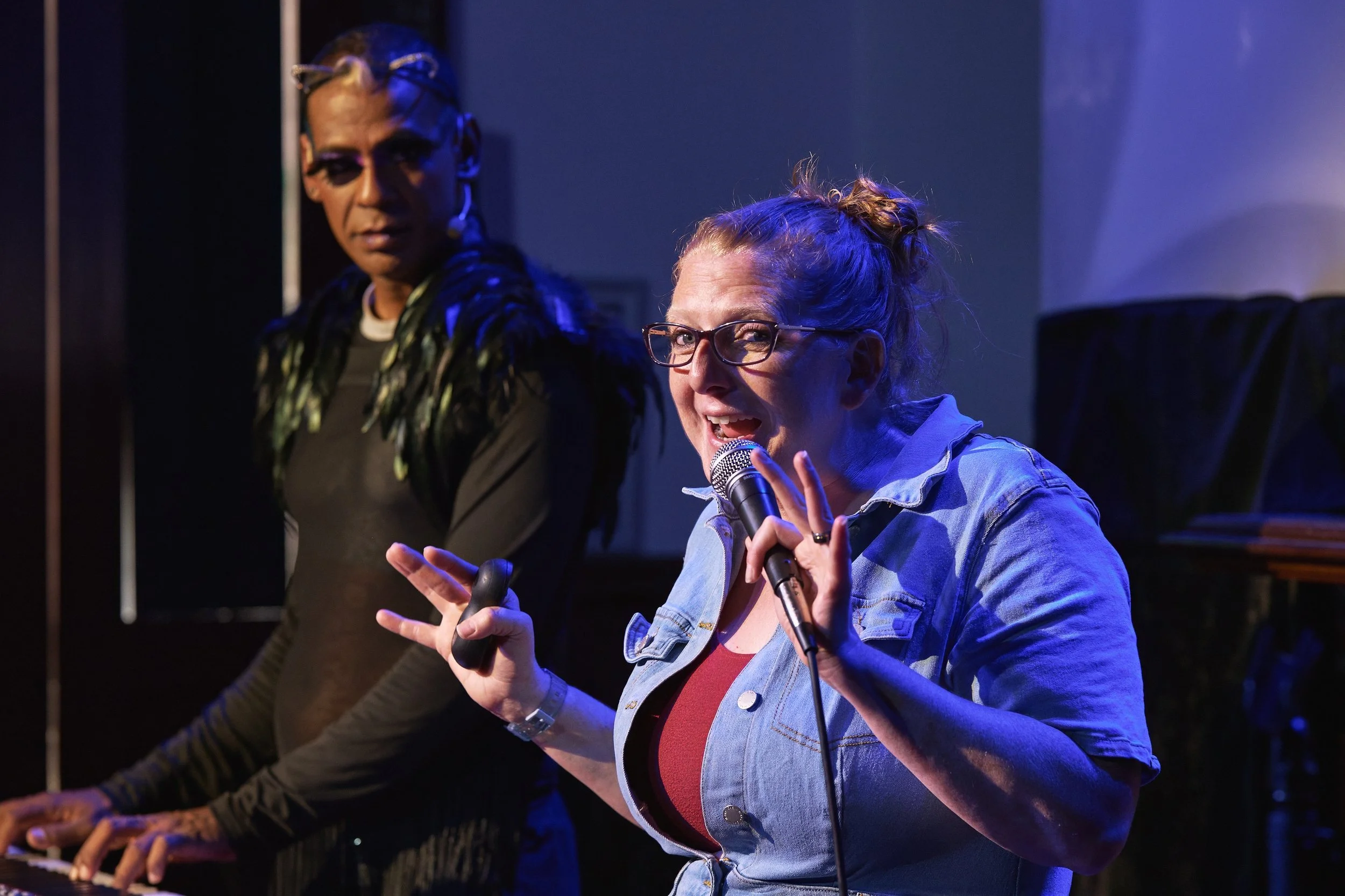 Comedian Robyn Perkins and Gavin Nicklette performing on stage, one singing into a microphone and the other playing keyboard, with colorful stage lighting.
