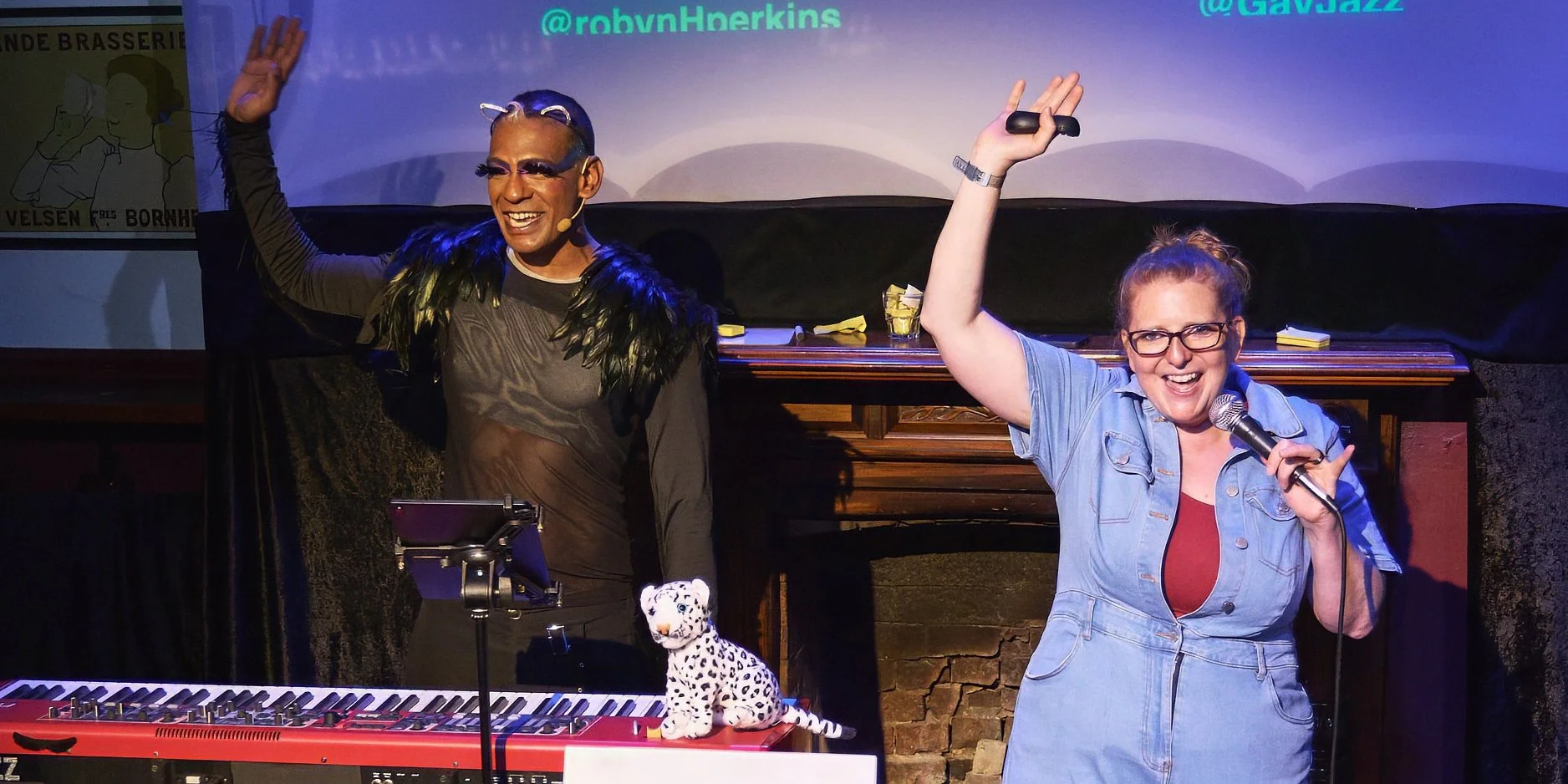 Comedian Robyn Perkins and Gavin Nicklette holding microphones and smiling and waving. A keyboard, a toy leopard, and a music stand are on the stage.