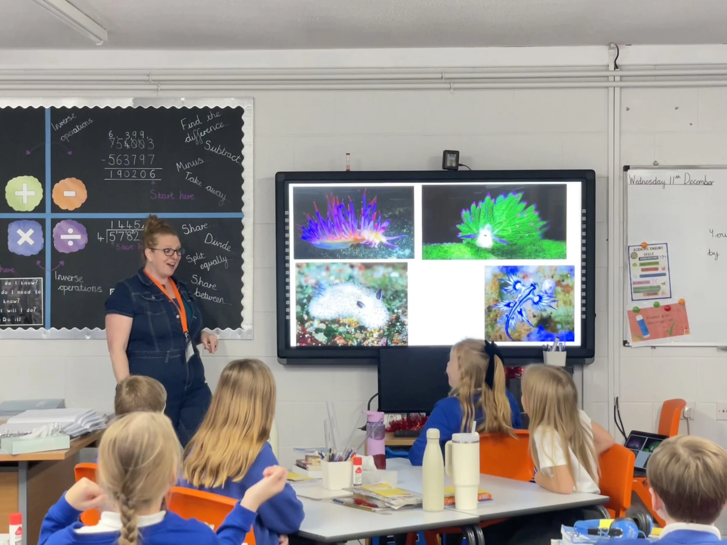 Comedian Robyn Perkins and young students in a classroom. The teacher stands near a digital display showing colorful images, while students seated at desks watch. Classroom walls feature educational posters and a whiteboard.