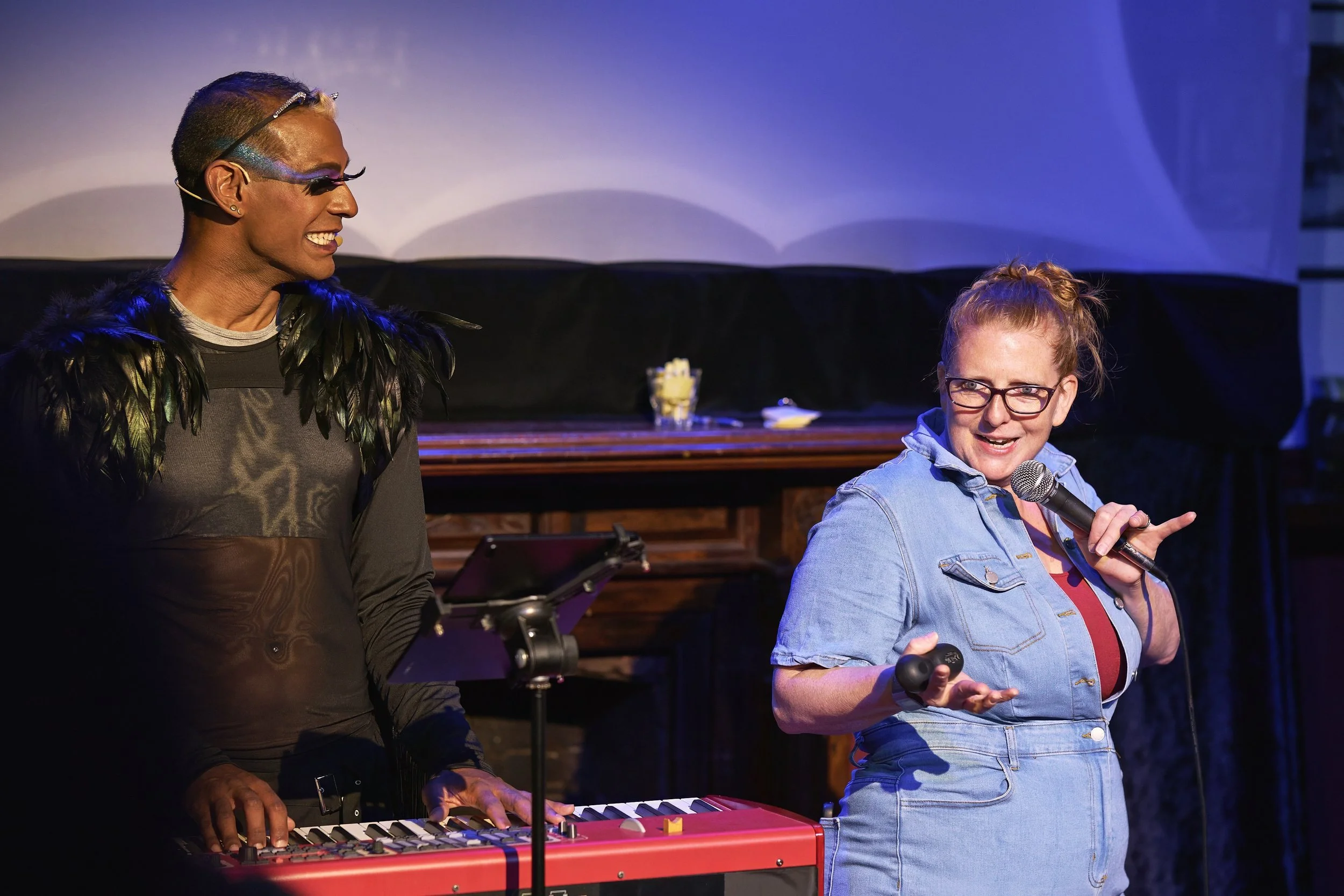 Comedian Robyn Perkins and Gavin Nicklette performing on stage; one playing a keyboard and dressed in black with feathered accessories, speaking into a microphone 