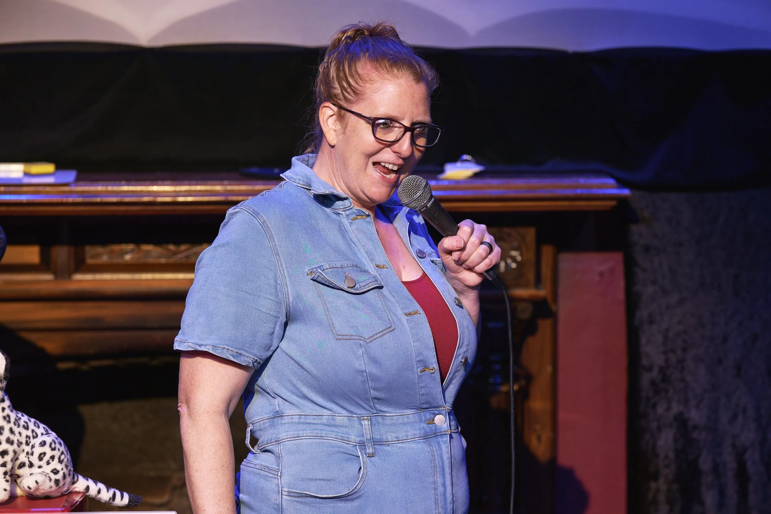 Comedian Robyn Perkins is holding a microphone and speaking on stage. There is a carved wooden piano behind her and a plush leopard toy on a table to her left.