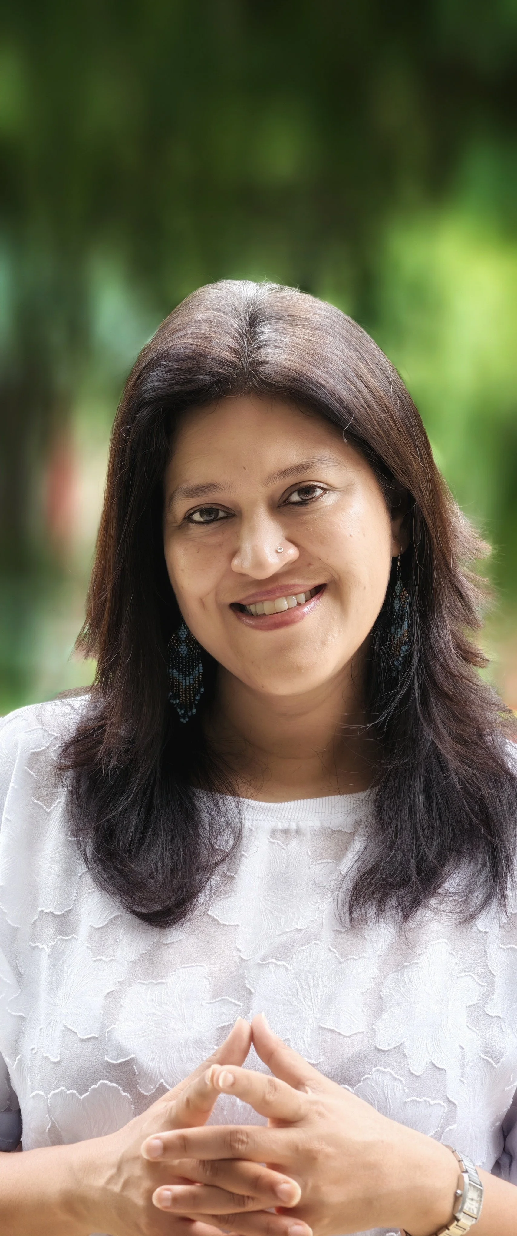 Smiling woman with dark brown hair, wearing earrings and a white textured top, posing outdoors with a blurred green forest background.