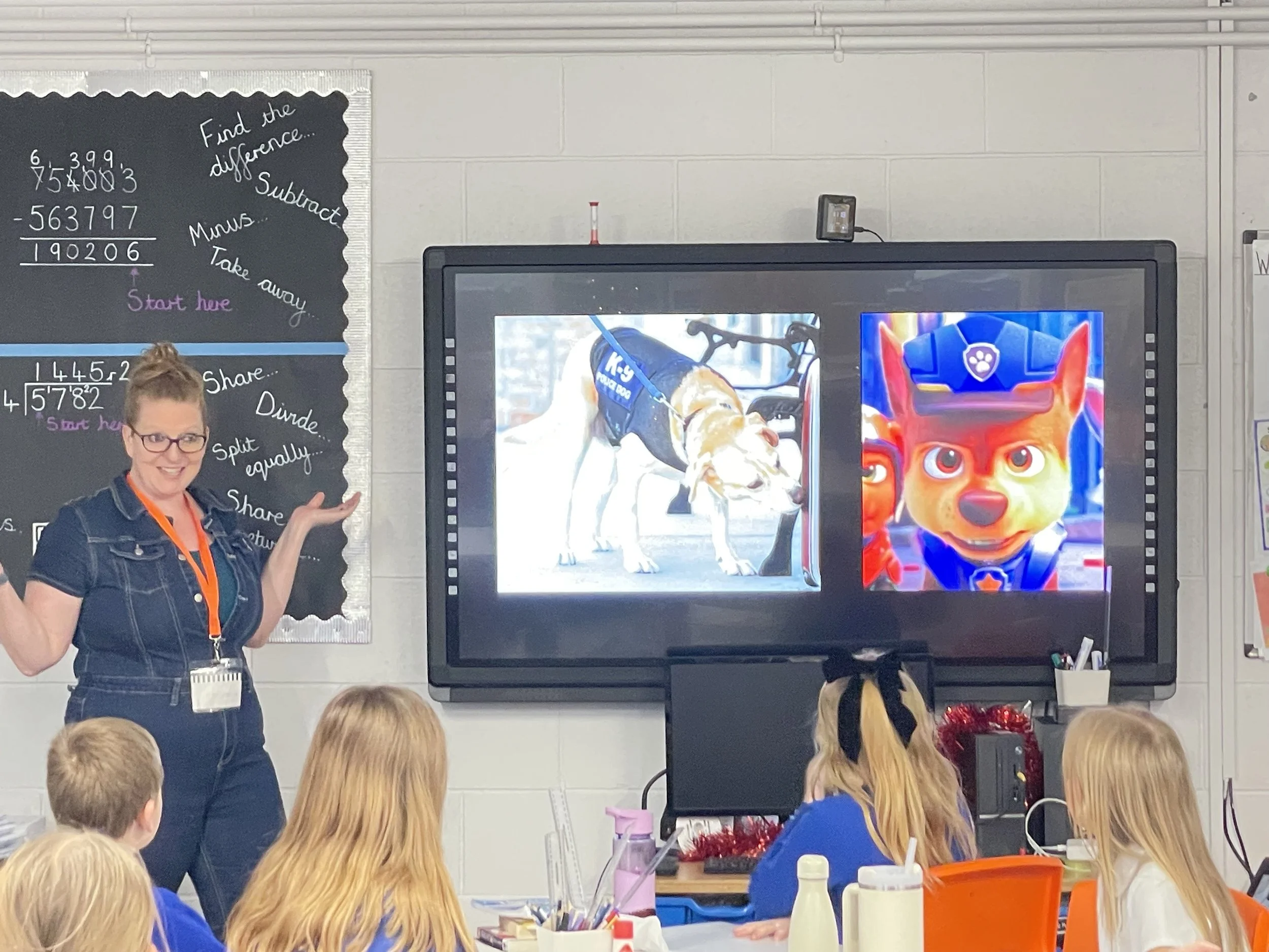 Comedian Robyn Perkins stands in front of a classroom pointing to two images on a digital screen. 