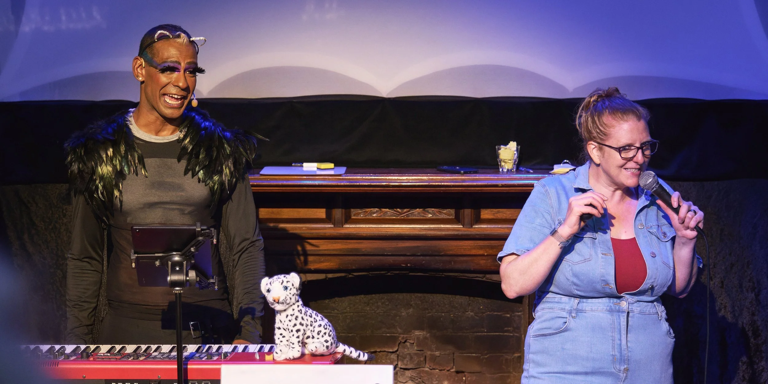 Comedian Robyn Perkins and Gavin Nicklette, smiling and standing by a keyboard with a stuffed white leopard toy holding a microphone and smiling. There are notes and a glass with a drink on stage.