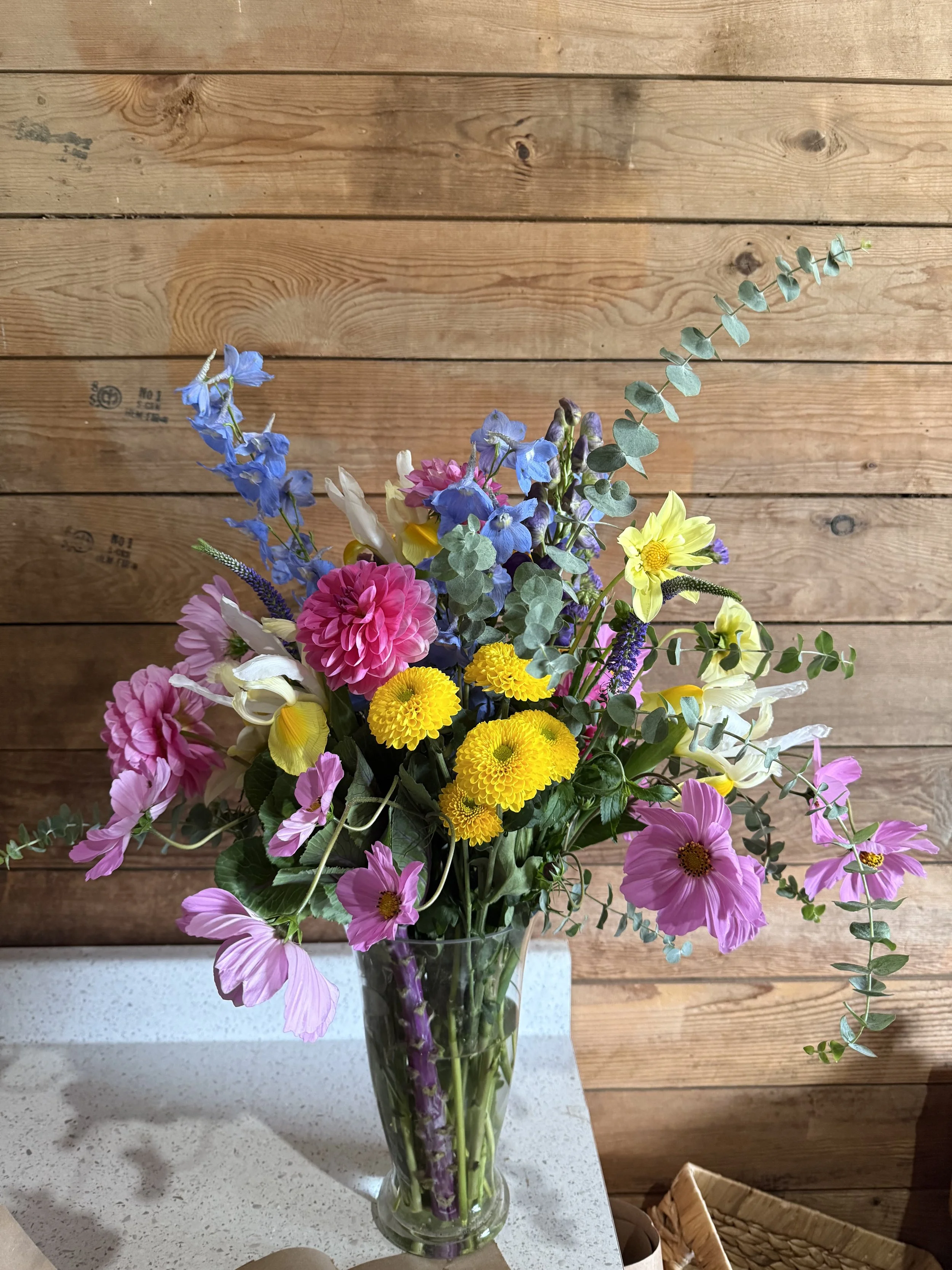 A colorful bouquet of mixed flowers in a glass vase on a white surface with a wooden wall background.