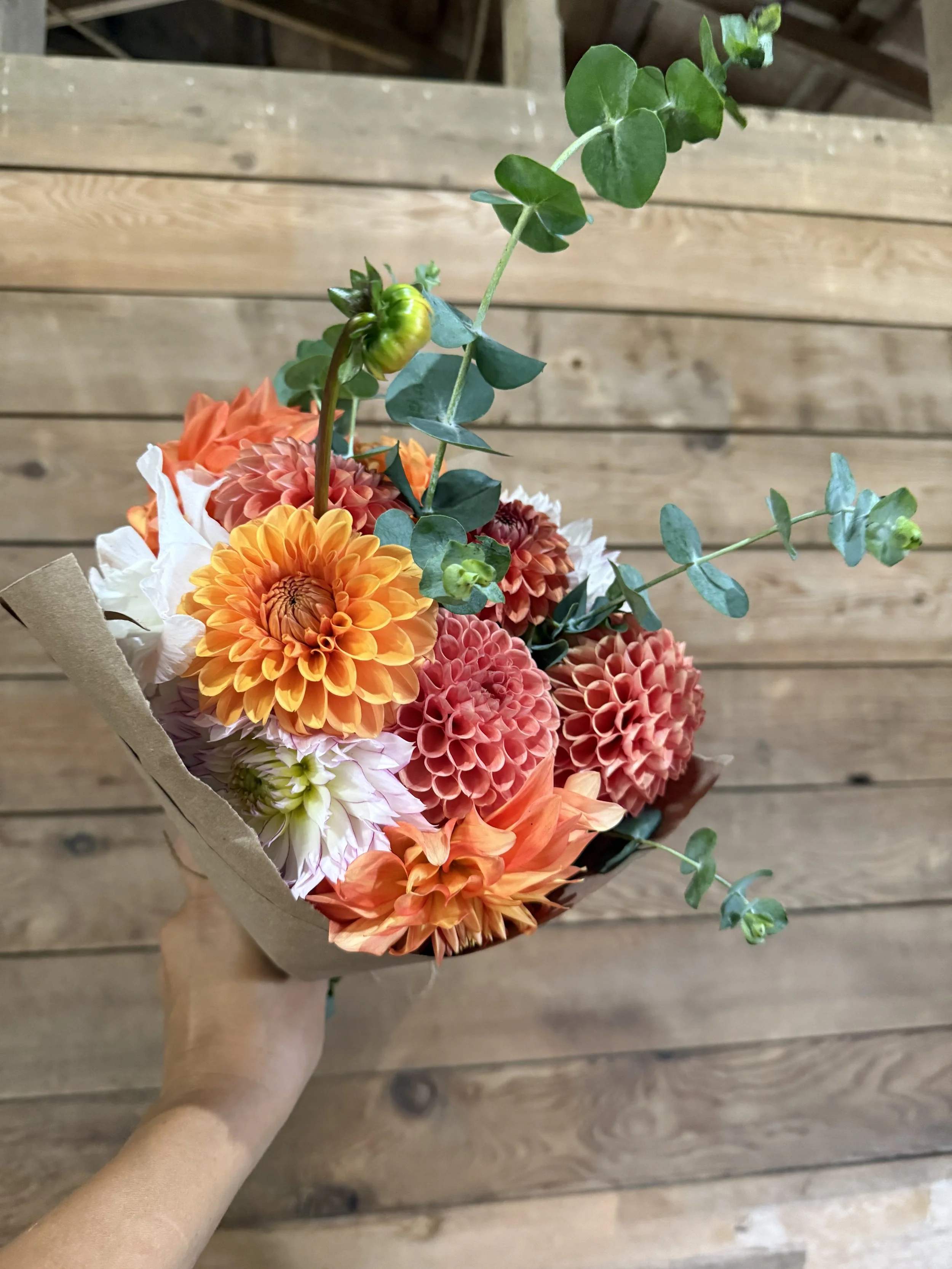 Hand holding a bouquet of orange, pink, and white flowers with green foliage on a wooden floor.