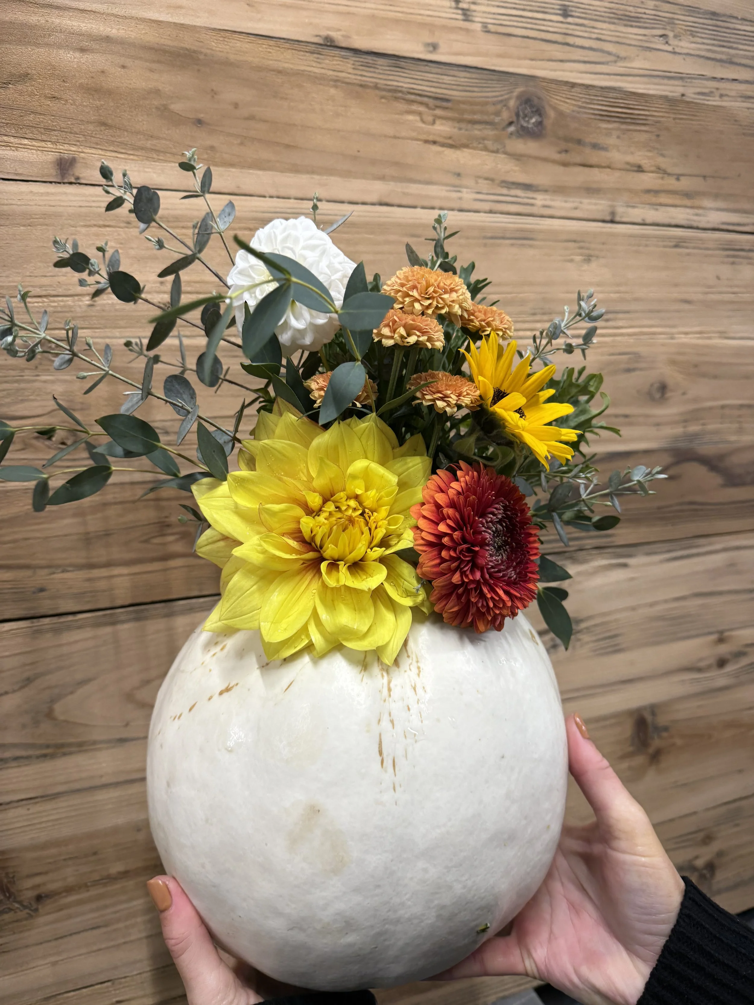 A person holding a white rounded ceramic vase filled with colorful flowers, including yellow, orange, red, and white blooms, with green foliage, against a wooden wall background.