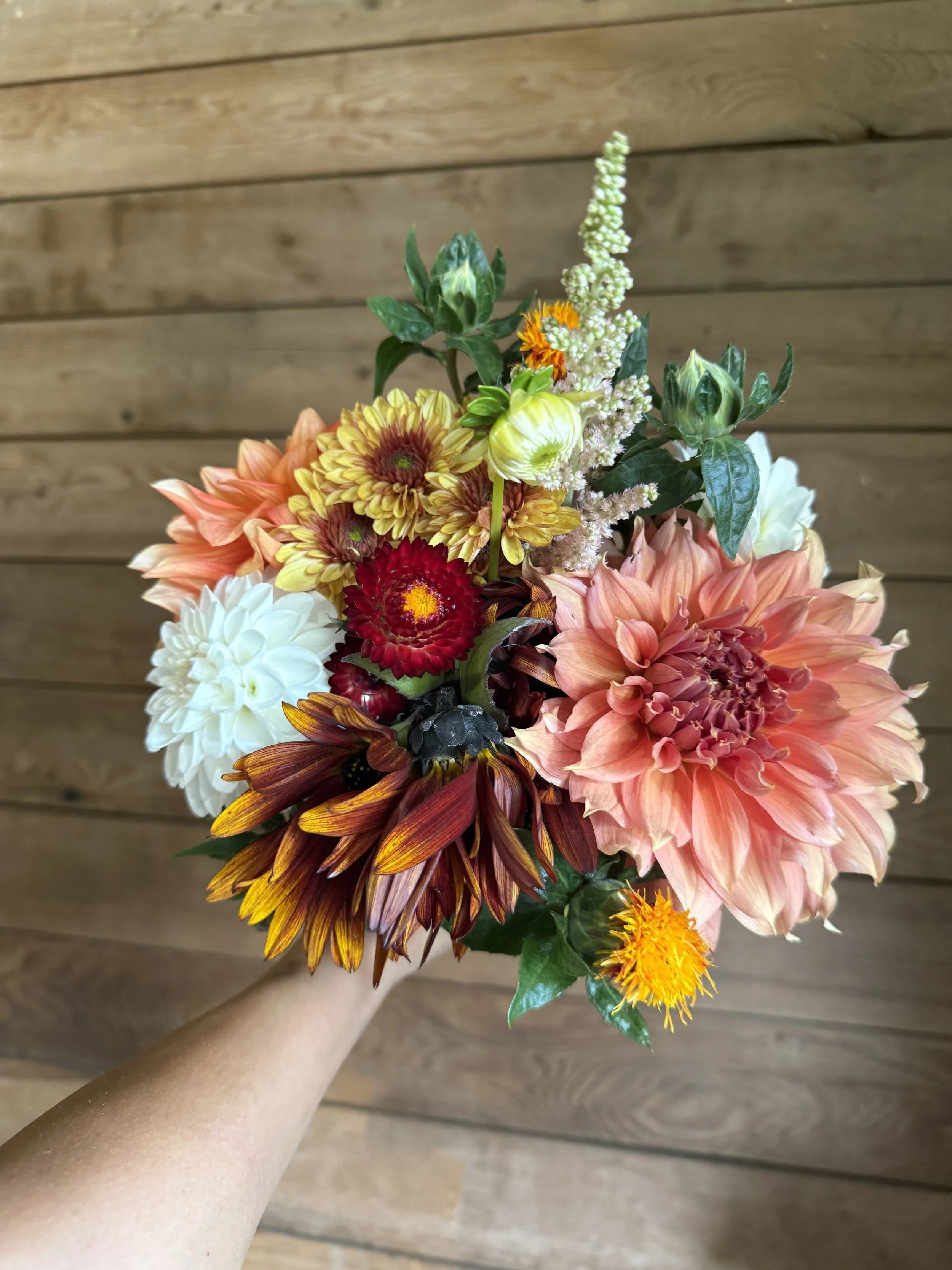 A colorful bouquet of various flowers including dahlias, chrysanthemums, and other blooms with green leaves, held in a person's hand against a wooden background.