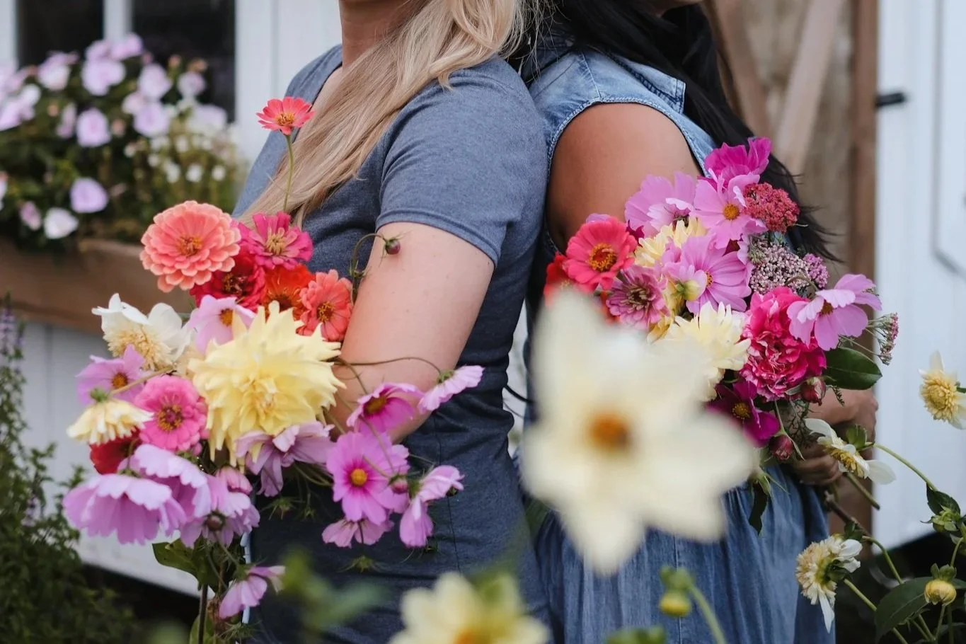 Two women standing back to back holding large bouquets of mixed pink, yellow, and white flowers.