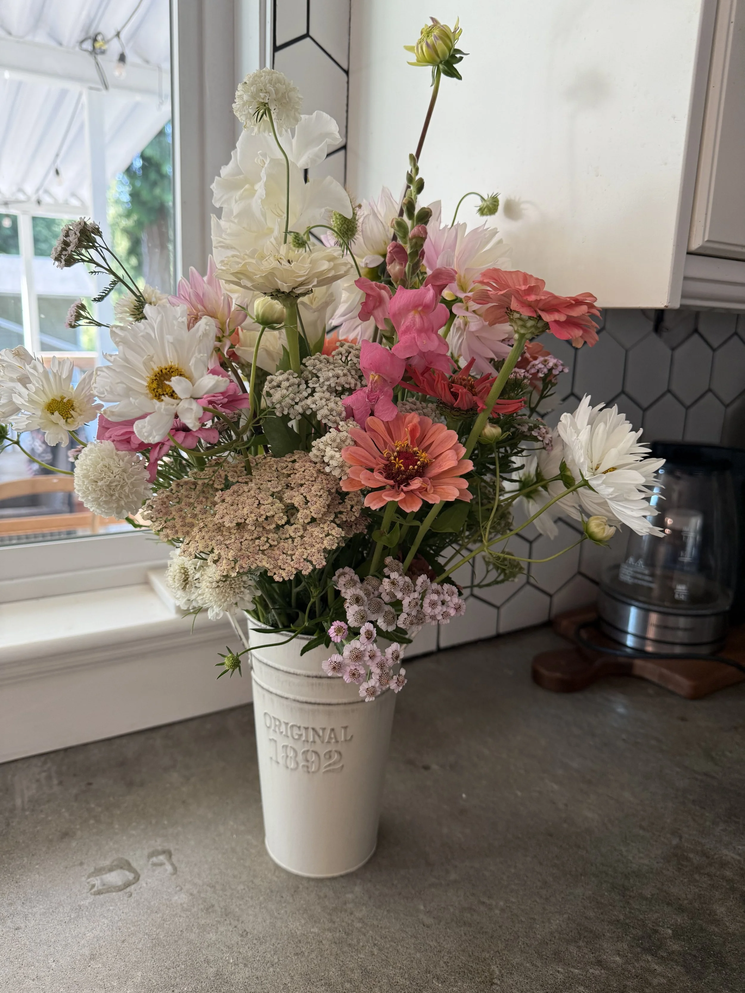 A bouquet of assorted flowers in a white vase on a kitchen countertop near a window, with a coffee maker in the background.