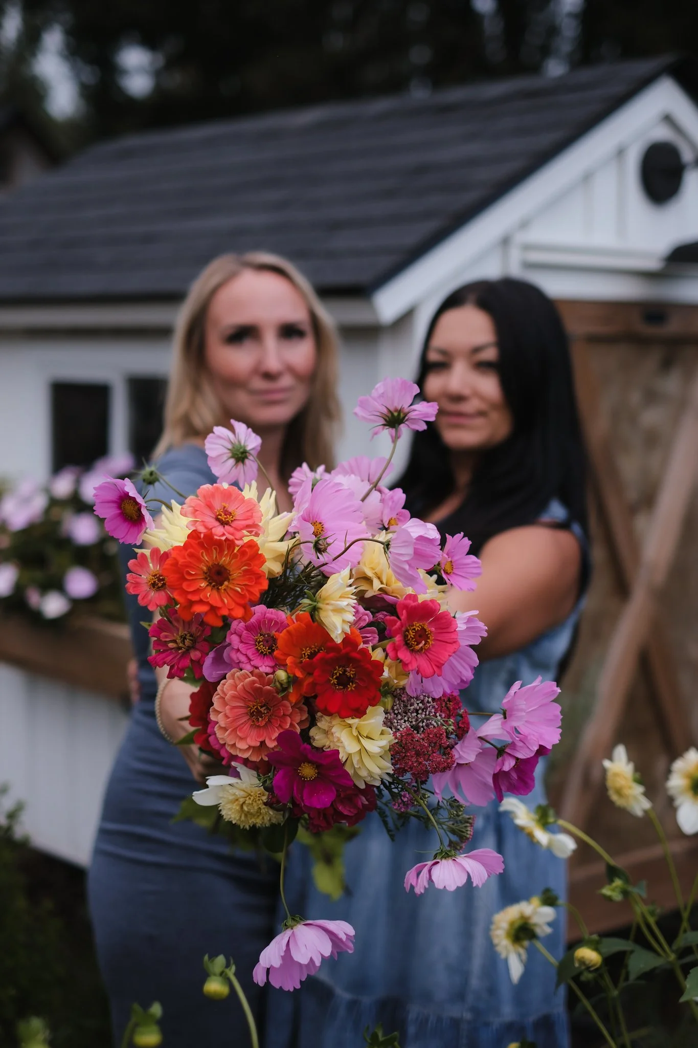 Two women hold a large colorful bouquet of flowers outdoors, with a small white house and garden in the background.