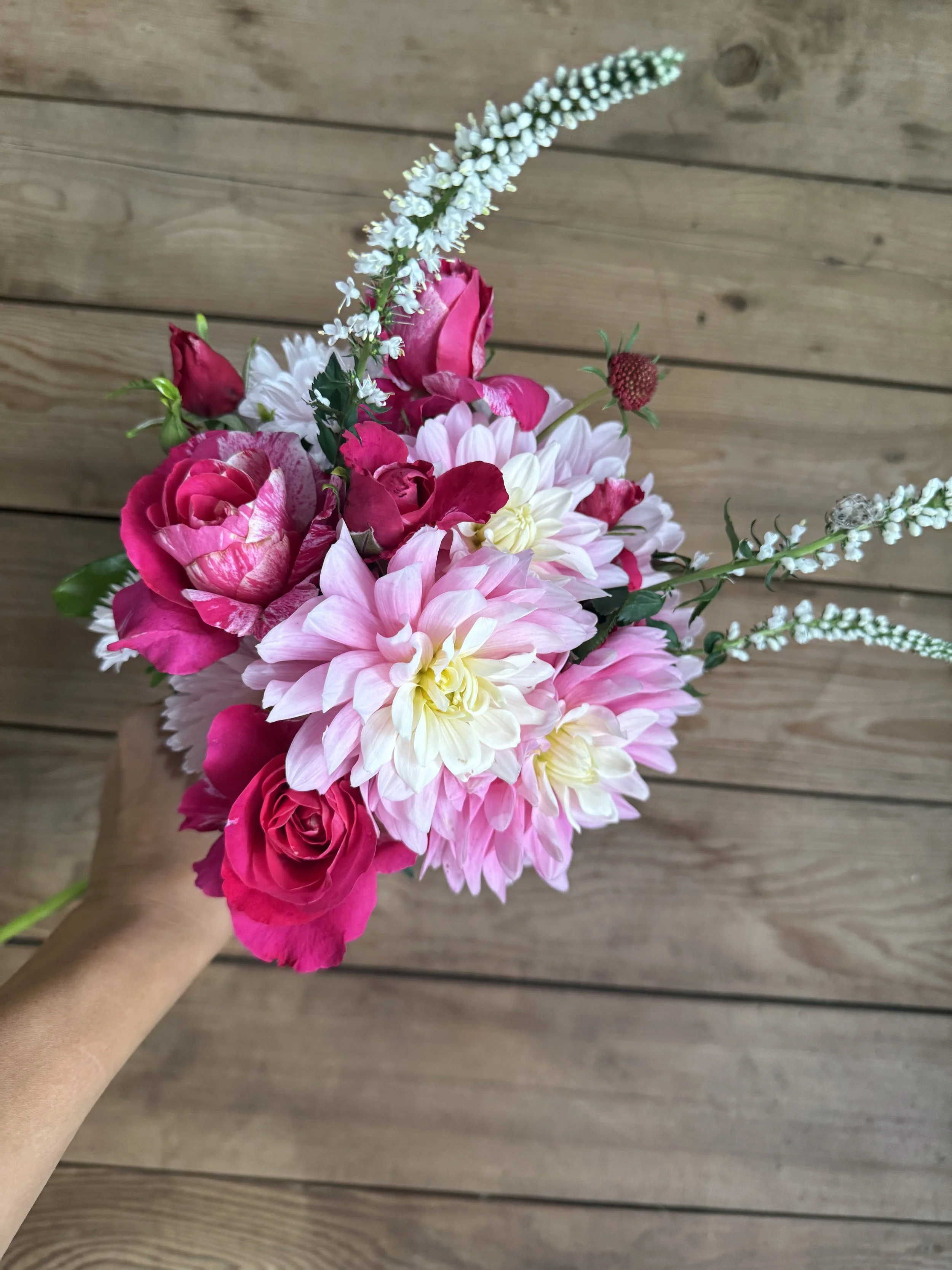 A colorful bouquet of pink and white flowers, including roses and dahlias, with white elongated flowers, against a wooden background.