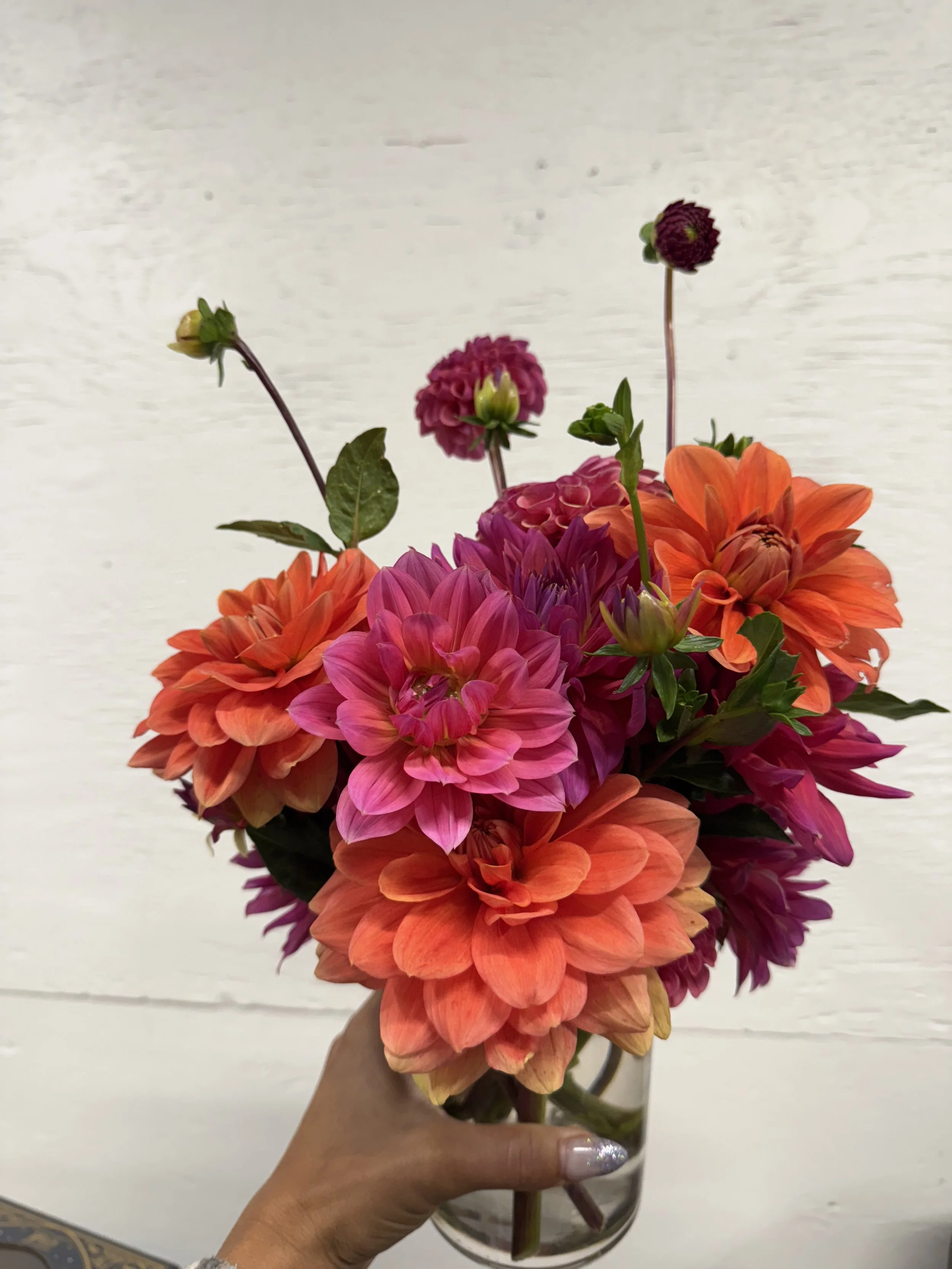 Hand holding a glass vase filled with colorful dahlias and other flowers against a white wooden background.