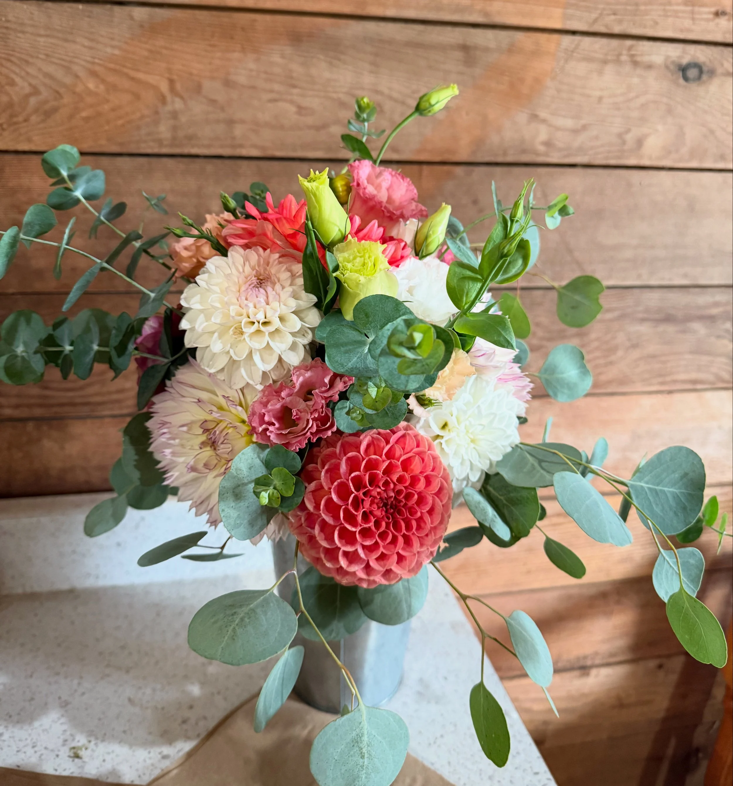 Colorful flower bouquet with dahlias, lisianthus, and eucalyptus leaves in a white vase placed on a table with a wooden wall background.