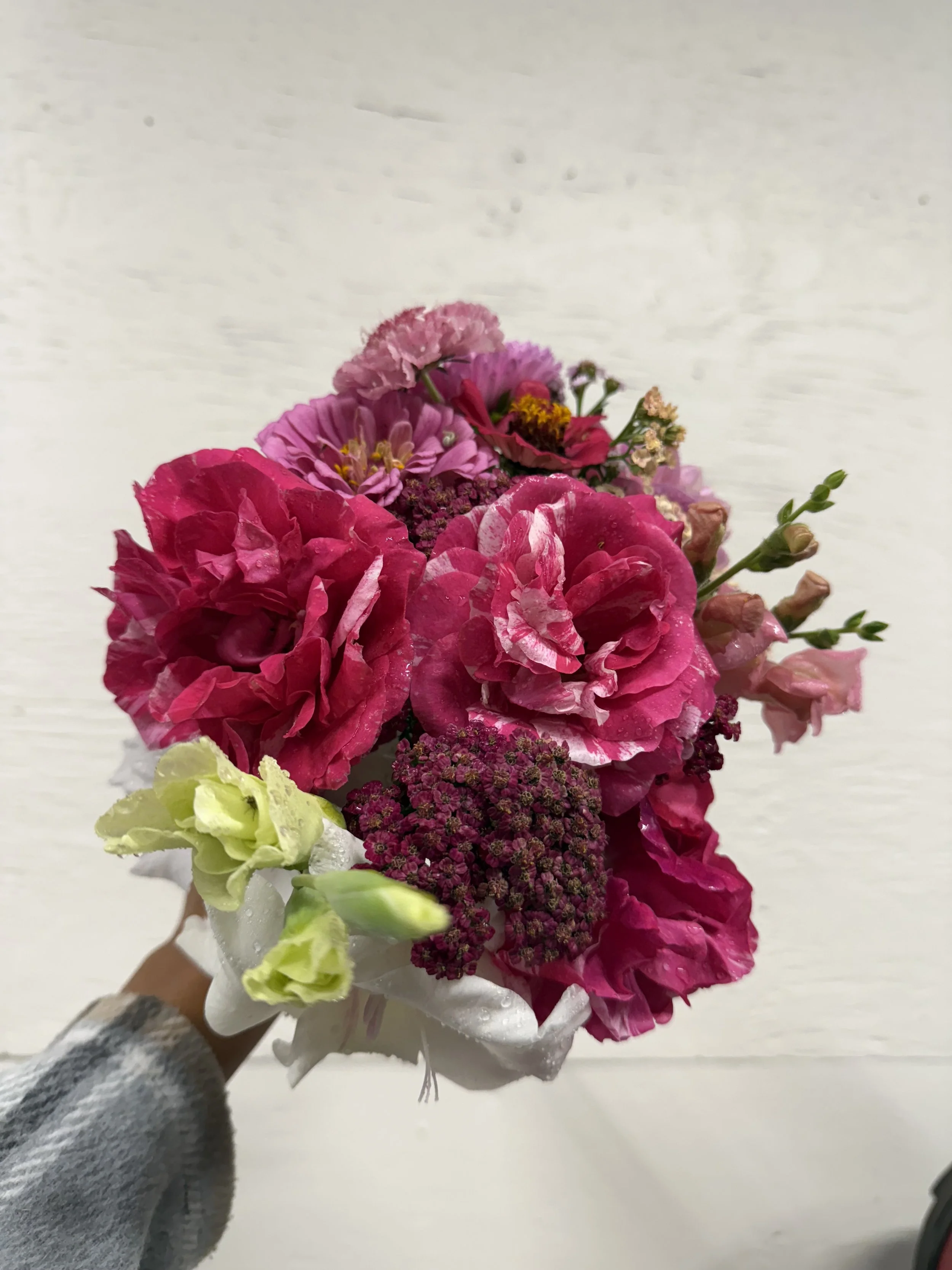 A hand holding a bouquet of pink, purple, and white flowers against a plain white background.