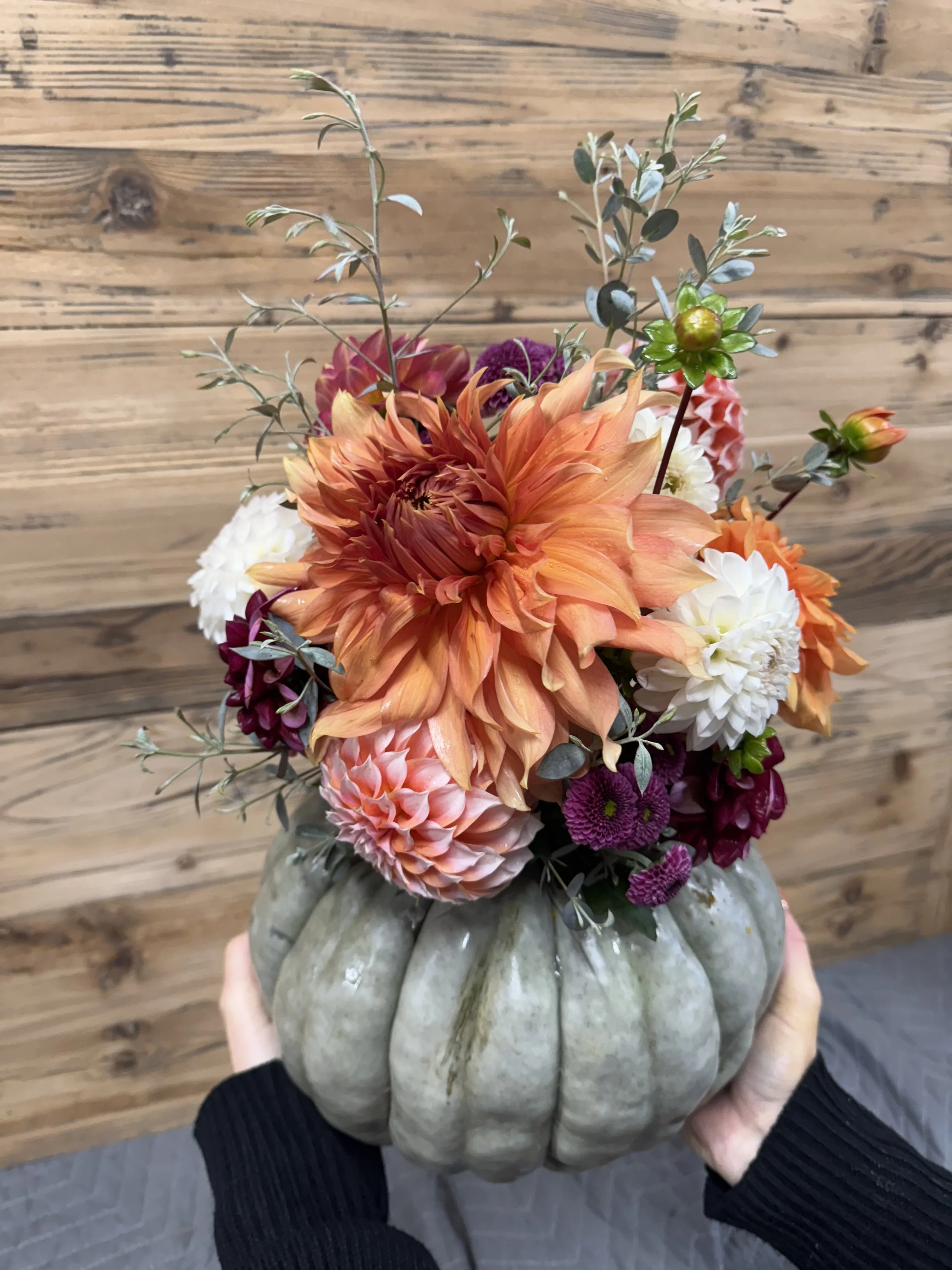 A person holding a decorative pumpkin-shaped vase filled with a colorful bouquet of flowers including orange dahlias, white dahlias, purple chrysanthemums, and green foliage against a wooden background.
